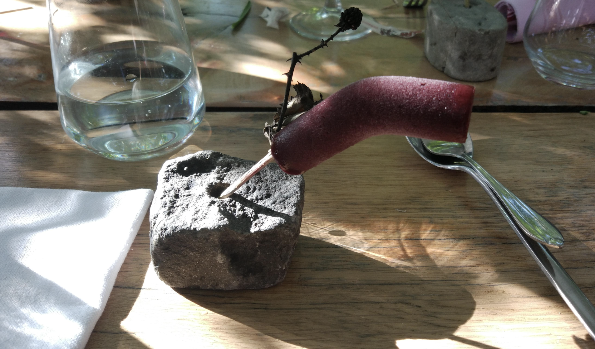 A purple popsicle on a stick is balanced in a hole in a gray rock, next to a glass of water, spoon, and napkin on a wooden table.