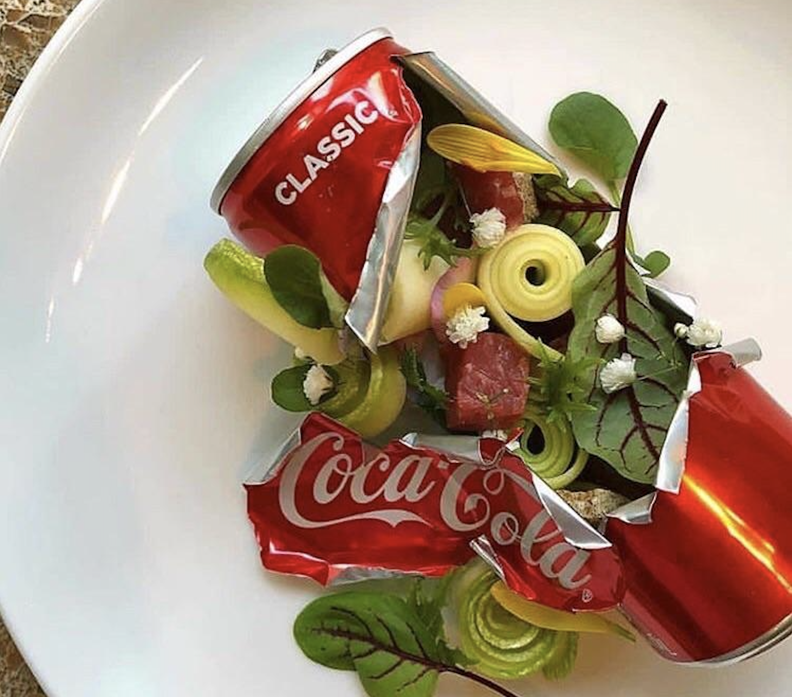 A deconstructed Coca-Cola can serves as a dish, filled with colorful vegetables, greens, and small cubes of meat, artfully plated on a white surface.