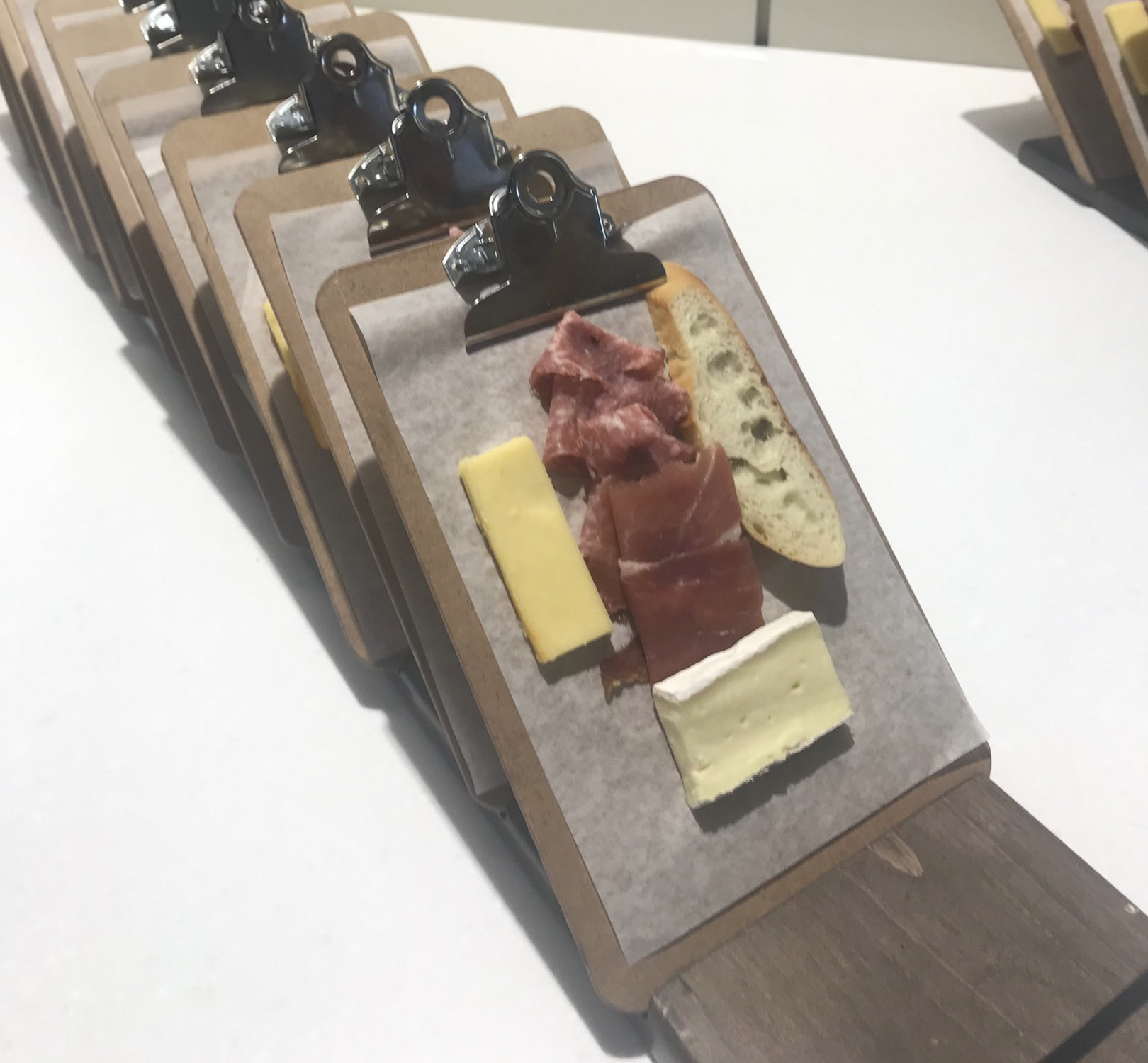 Small clipboards display appetizers of sliced cheese, cured meat, and a piece of bread, arranged neatly in a row on white parchment paper.