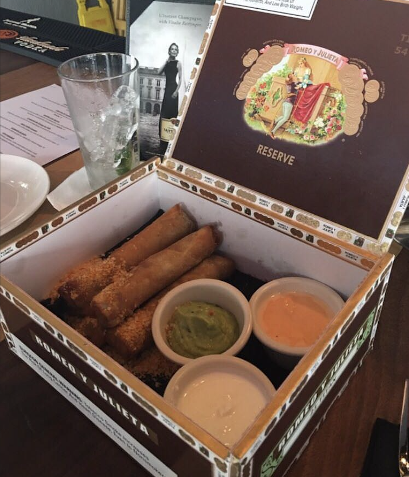 Four crispy spring rolls and three dipping sauces are served in a repurposed cigar box on a restaurant table, with a glass of iced drink and menus visible in the background.