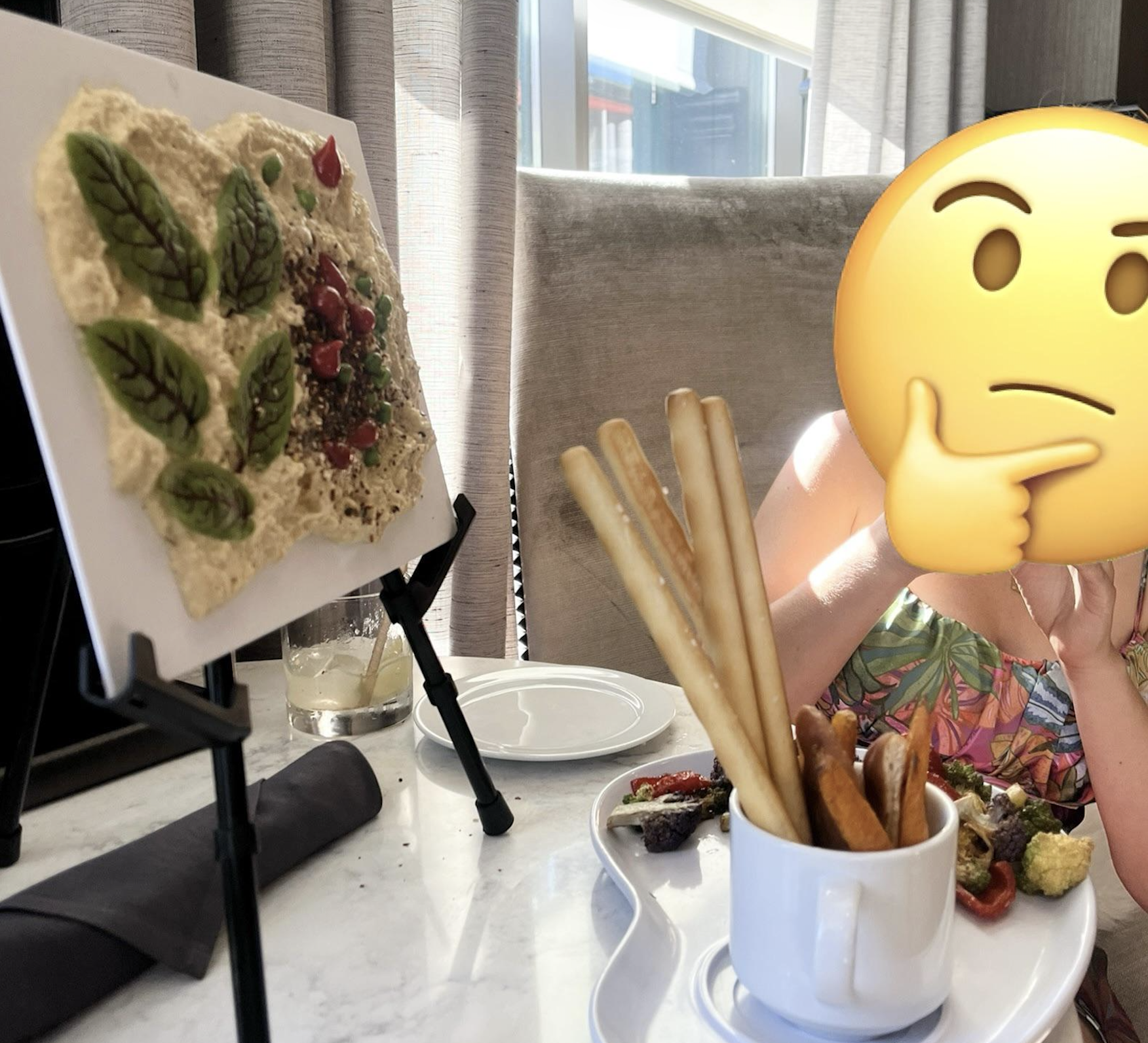 A person with a thinking face emoji sits at a table with a plate of breadsticks and snacks. Next to them, a cracker with leaf decorations is displayed upright on an easel. Sunlight streams through a window behind.
