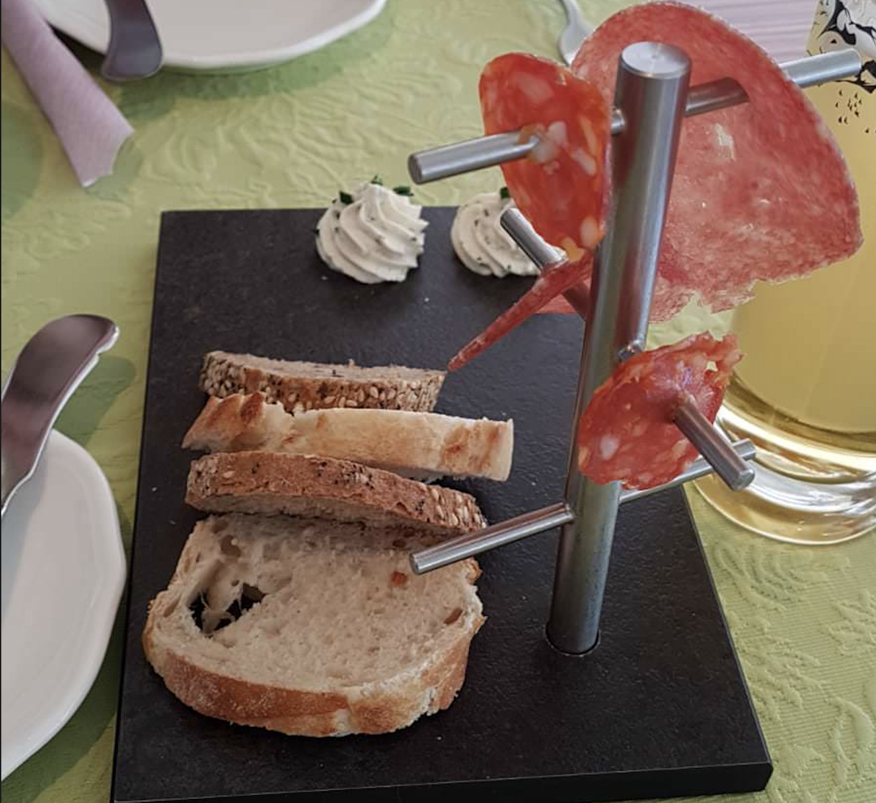 Slices of bread stacked on a black serving board next to a metal stand holding slices of salami, with two swirls of herb butter and a glass of light beverage in the background.