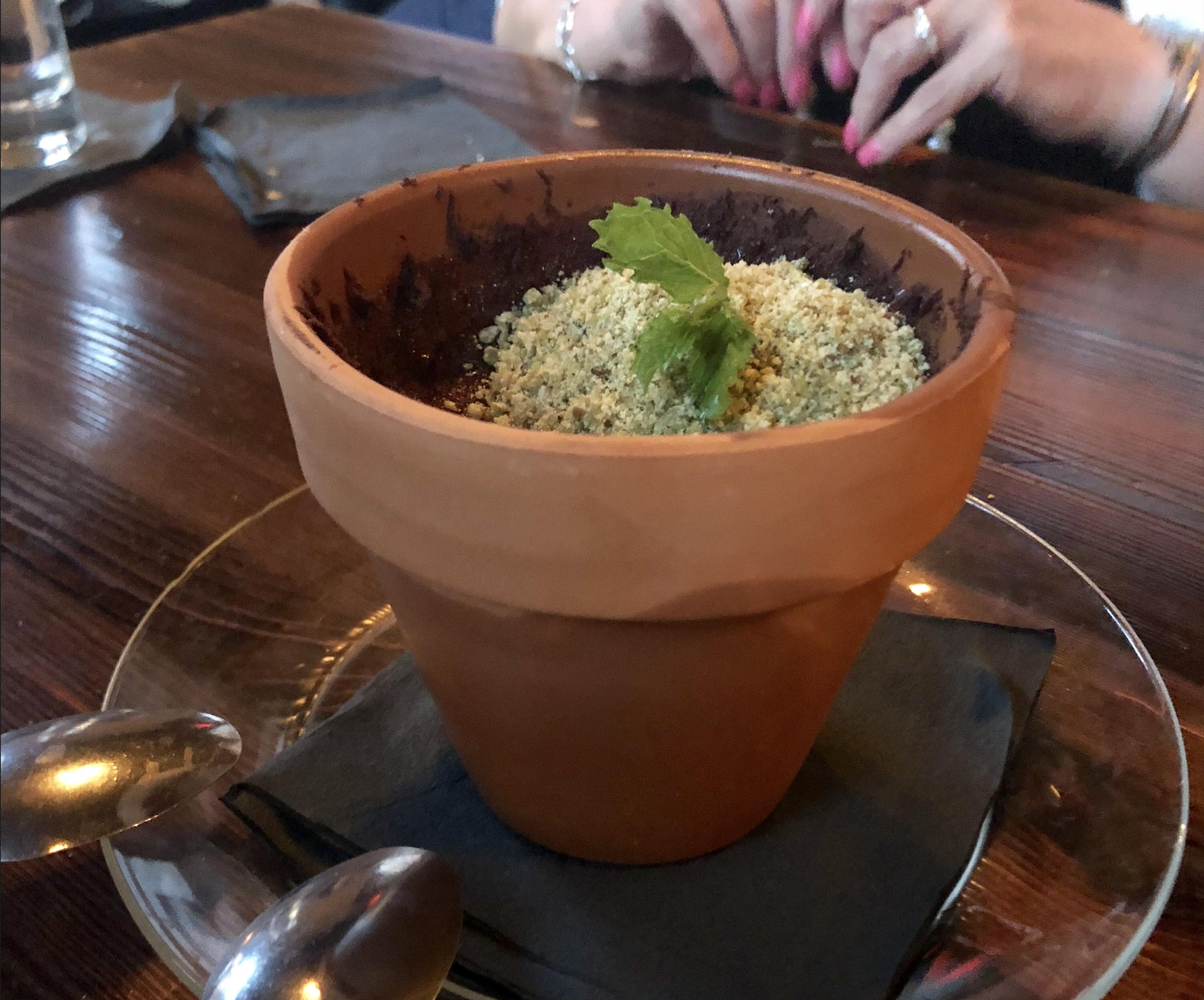 A dessert served in a small flowerpot, topped with crumbled cookie “soil” and a mint leaf, placed on a napkin with spoons nearby. A person’s hands can be seen in the background.