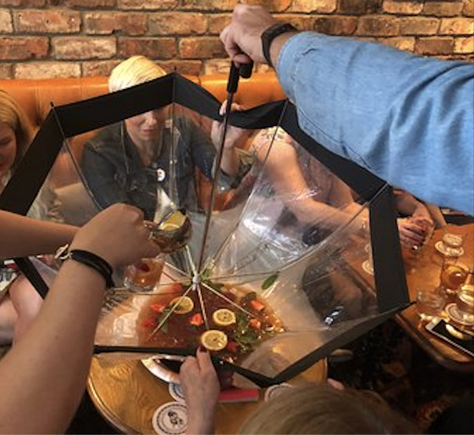 Several people sit around a table using an upside-down umbrella as a bowl, filling it with drinks, lemon slices, and fruit in a lively indoor setting.