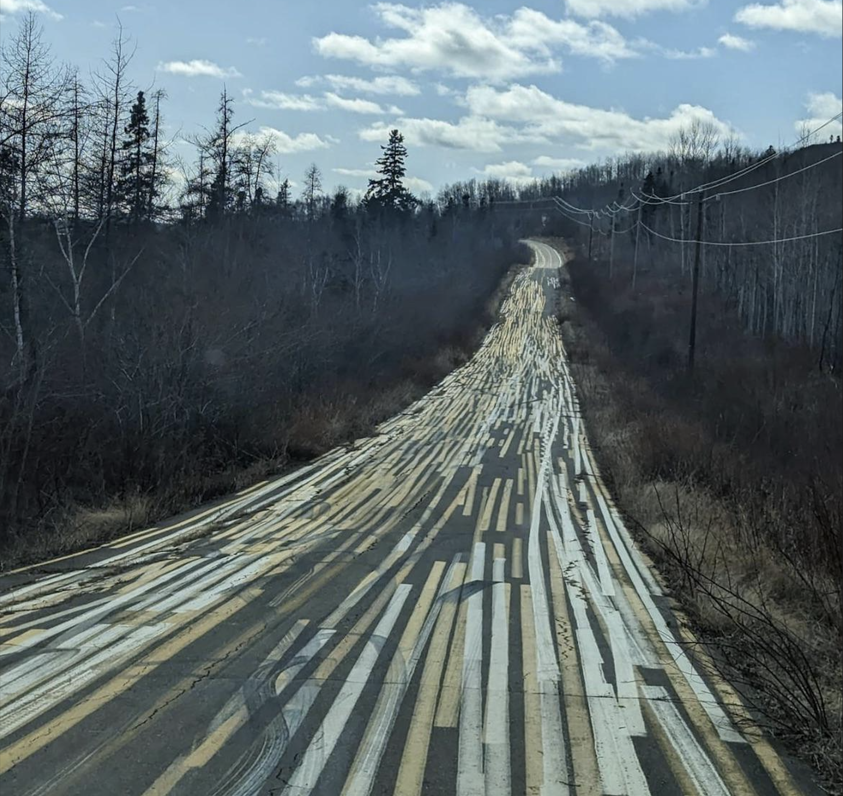 A rural road with unusually striped, patchy repairs stretches into the distance, surrounded by leafless trees under a partly cloudy sky.