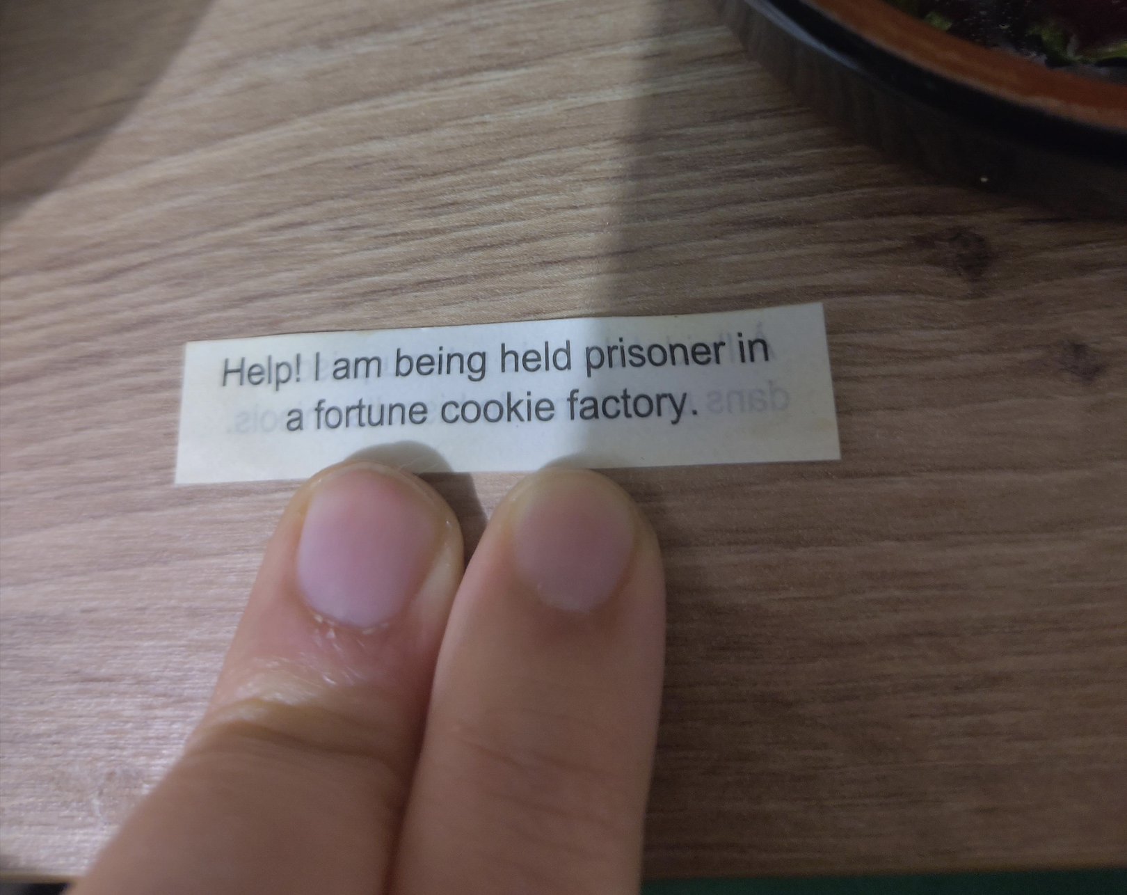 A hand holds a fortune slip that reads, “Help! I am being held prisoner in a fortune cookie factory,” on a wooden surface.