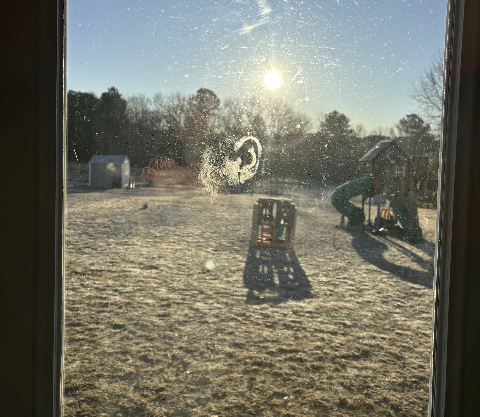 A frosty backyard is seen through a window with a nose and partial face print. The sun is low, casting long shadows over playground equipment and a shed on the grassy lawn.