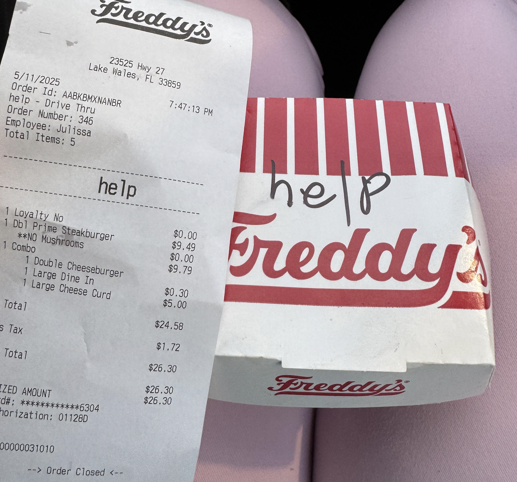 A Freddy’s receipt and food box on someone’s pink-clad lap. The word “help” is handwritten on the food box lid above the Freddy’s logo.