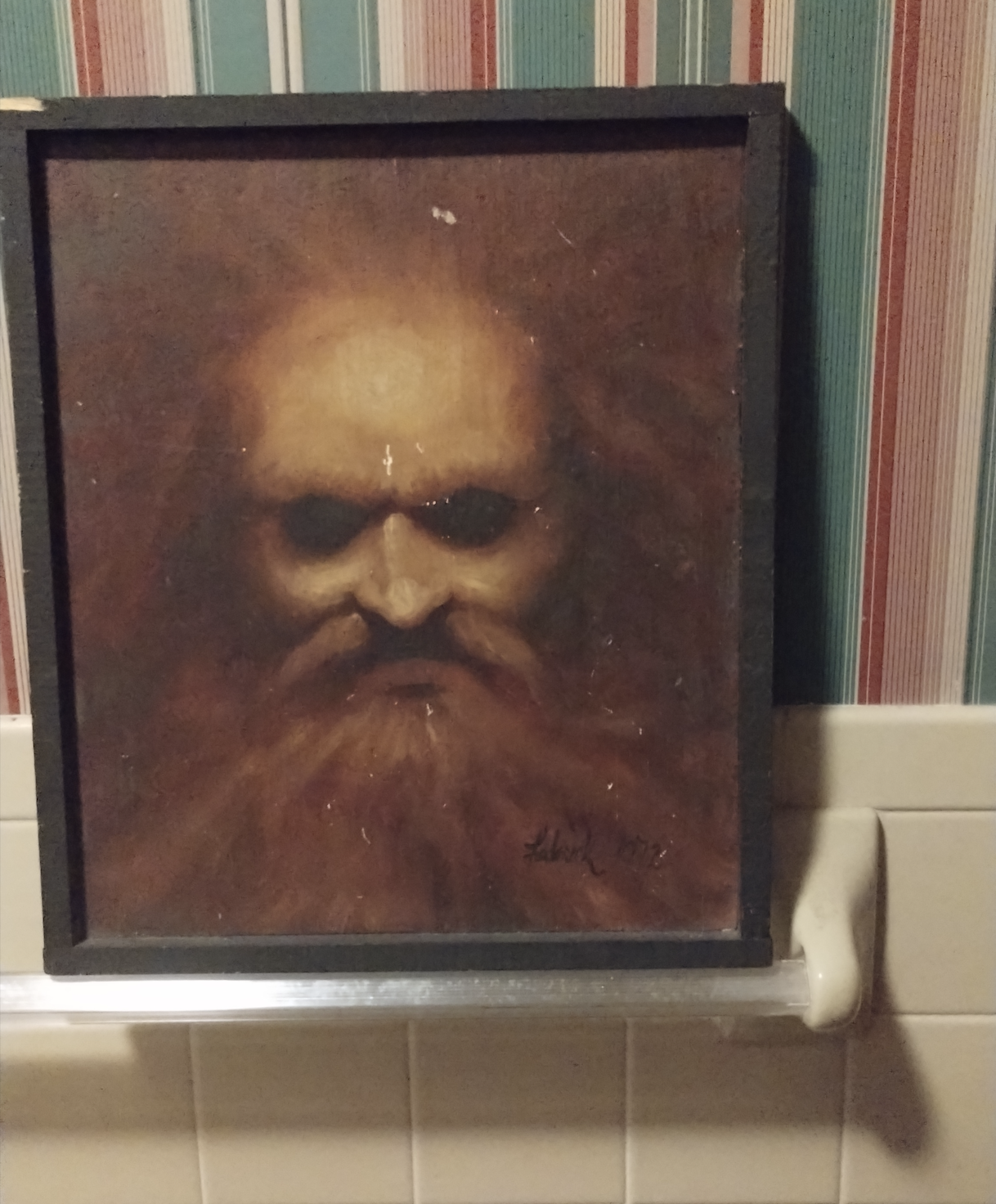 A framed painting of a bearded face with intense eyes and shadowy lighting hangs on a bathroom wall with striped wallpaper and white tiles.