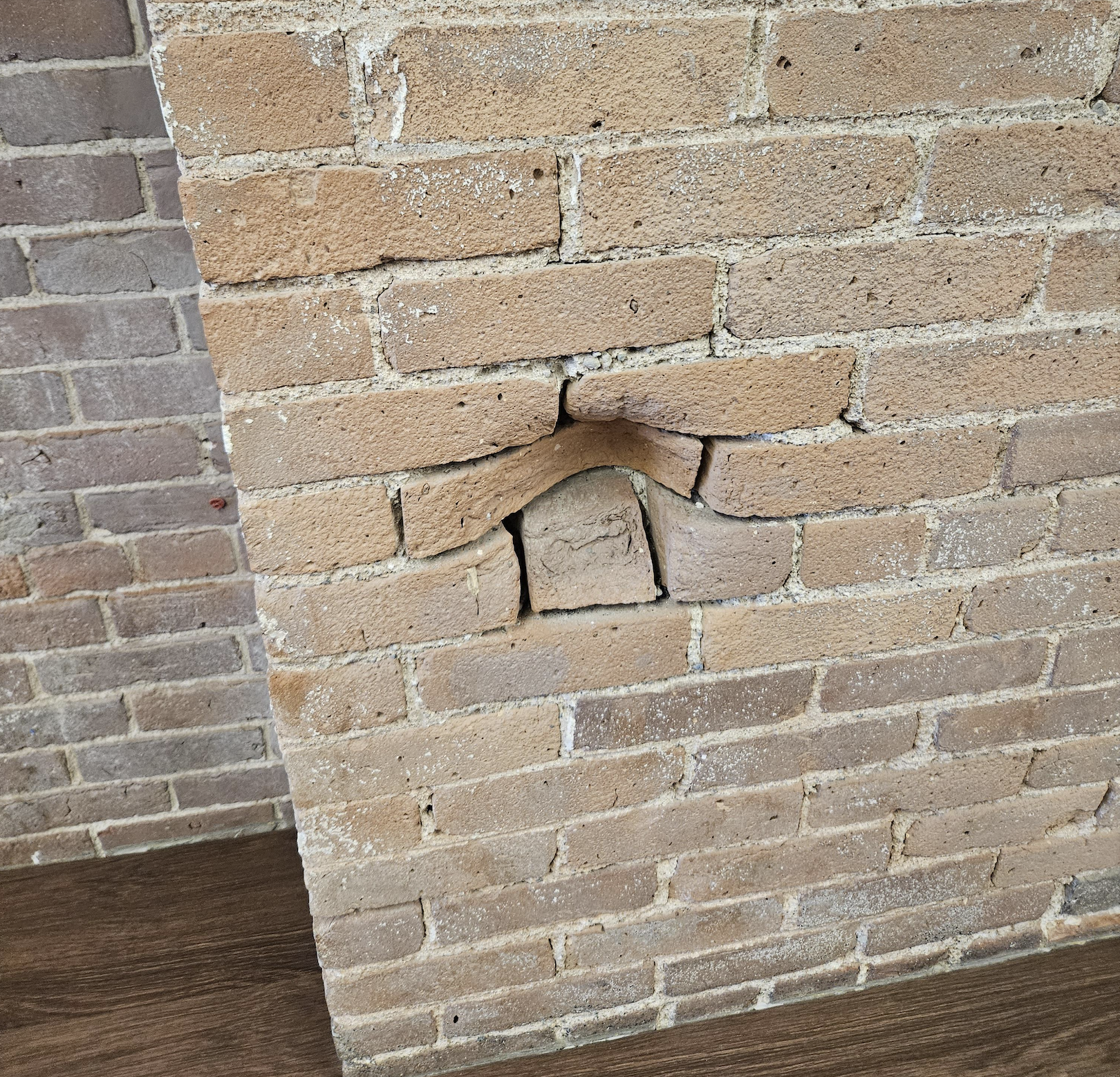 A close-up of a tan brick wall with several bricks oddly arranged, forming a small arch-like bump protruding outward above a patch of mismatched mortar. Brown wooden floor is visible at the bottom.