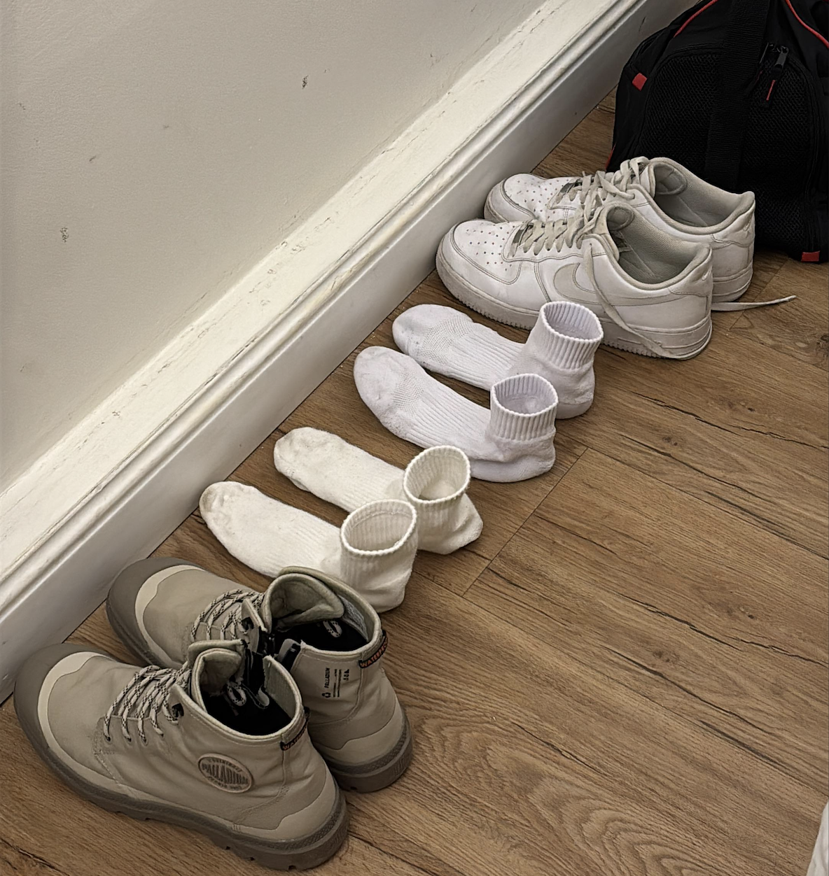 Three pairs of white socks neatly placed in between a pair of beige boots and a pair of white sneakers, lined up against a white wall on a wooden floor. A black bag is in the background.