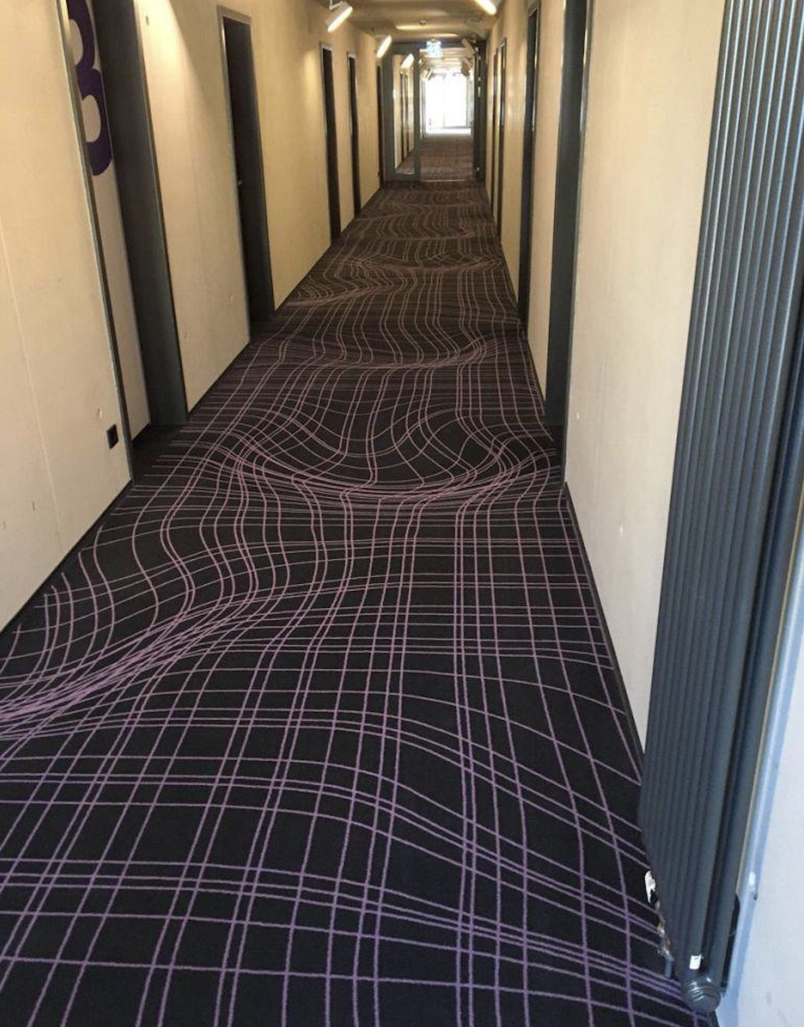 A long hallway with a carpet featuring wavy, purple and pink grid lines that create an optical illusion, making the floor appear uneven and distorted.