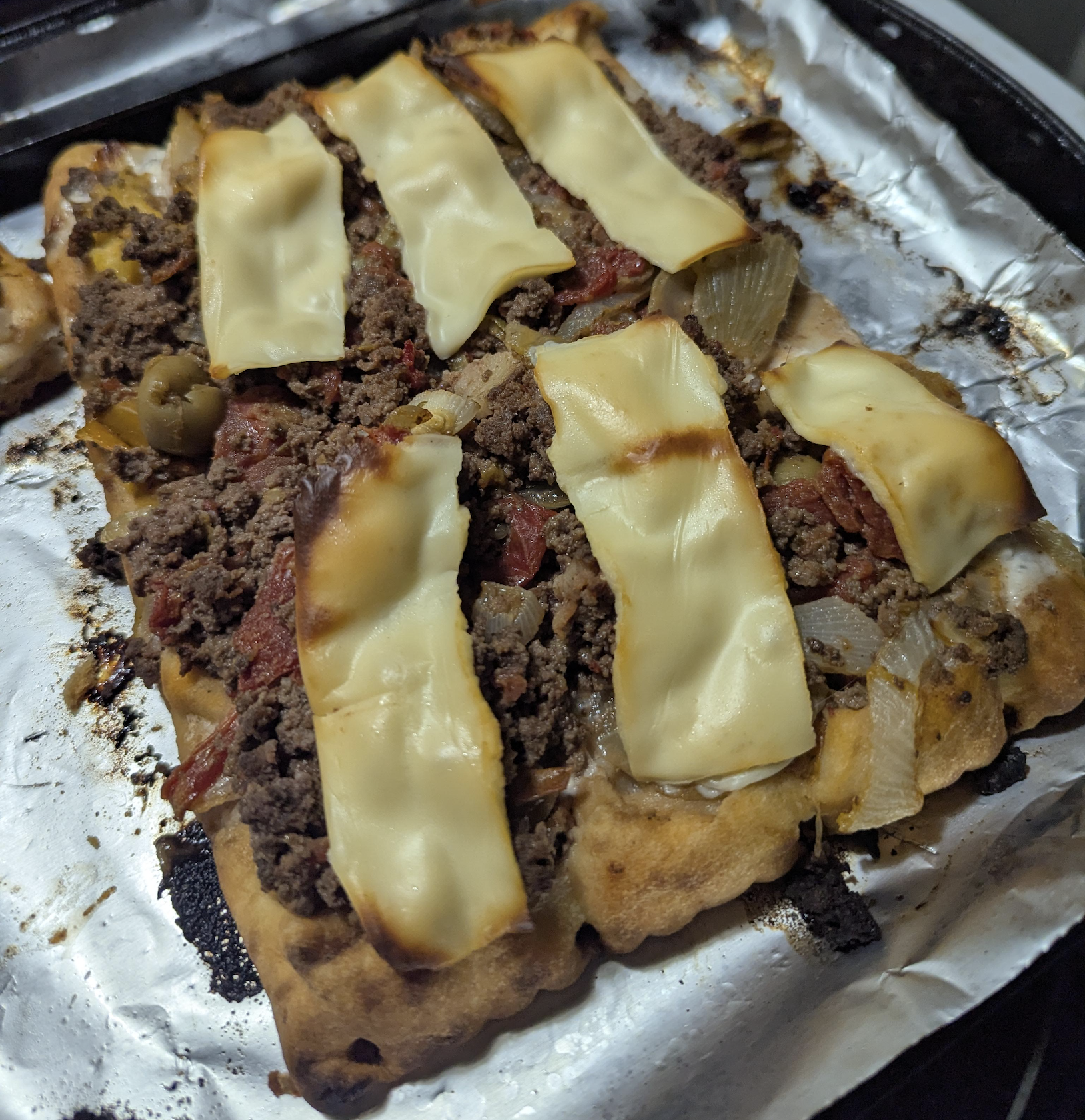 A rectangular flatbread topped with ground beef, onions, olives, tomatoes, and melted slices of cheese, baked on a foil-lined pan.