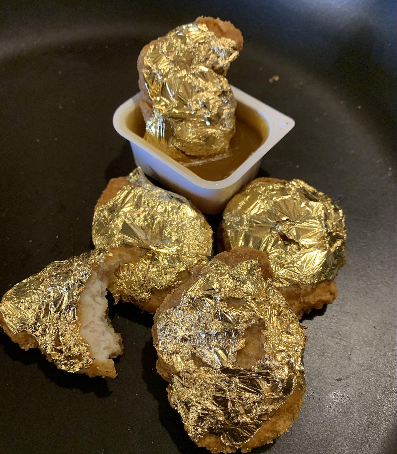 Four chicken nuggets covered in gold leaf are arranged around a dipping sauce container, with one nugget partially dipped and another showing a bite taken out of it.