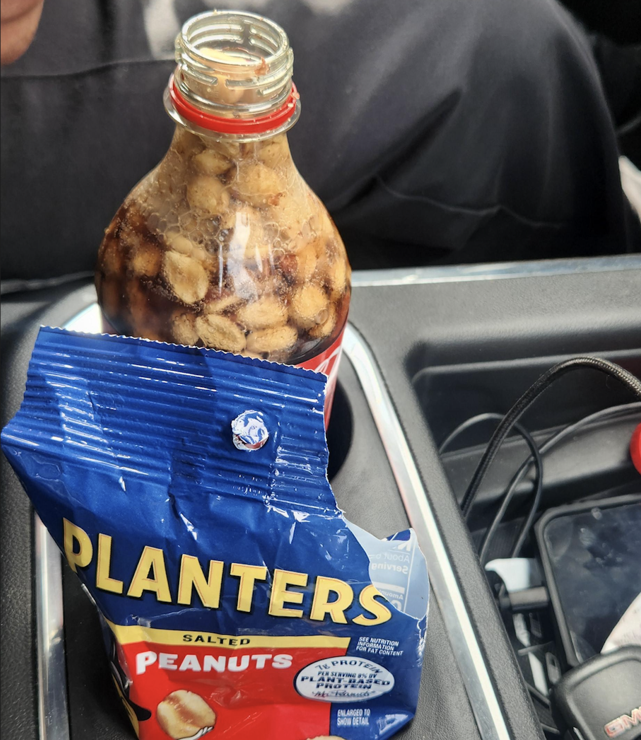 An open bottle of cola with peanuts inside sits in a car cupholder next to an opened blue bag of Planters salted peanuts.