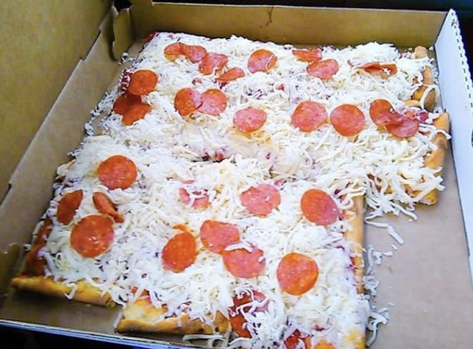 A rectangular pizza in a box with unevenly melted cheese and pepperoni slices, appearing undercooked and unfinished. Some cheese is not melted and scattered loosely.