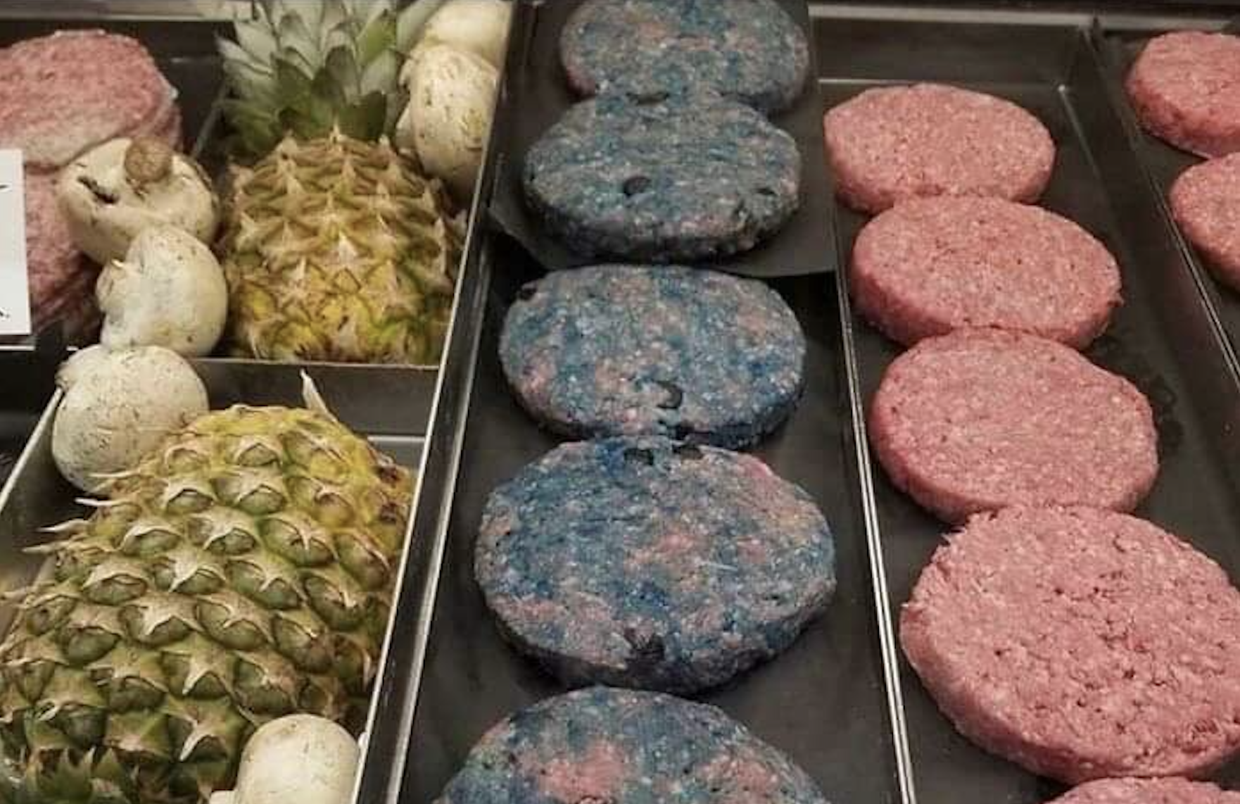 Raw hamburger patties are displayed in a tray, with one row of blue-and-pink patties next to a row of plain pink ones. Pineapples and mushrooms are also visible beside the trays.