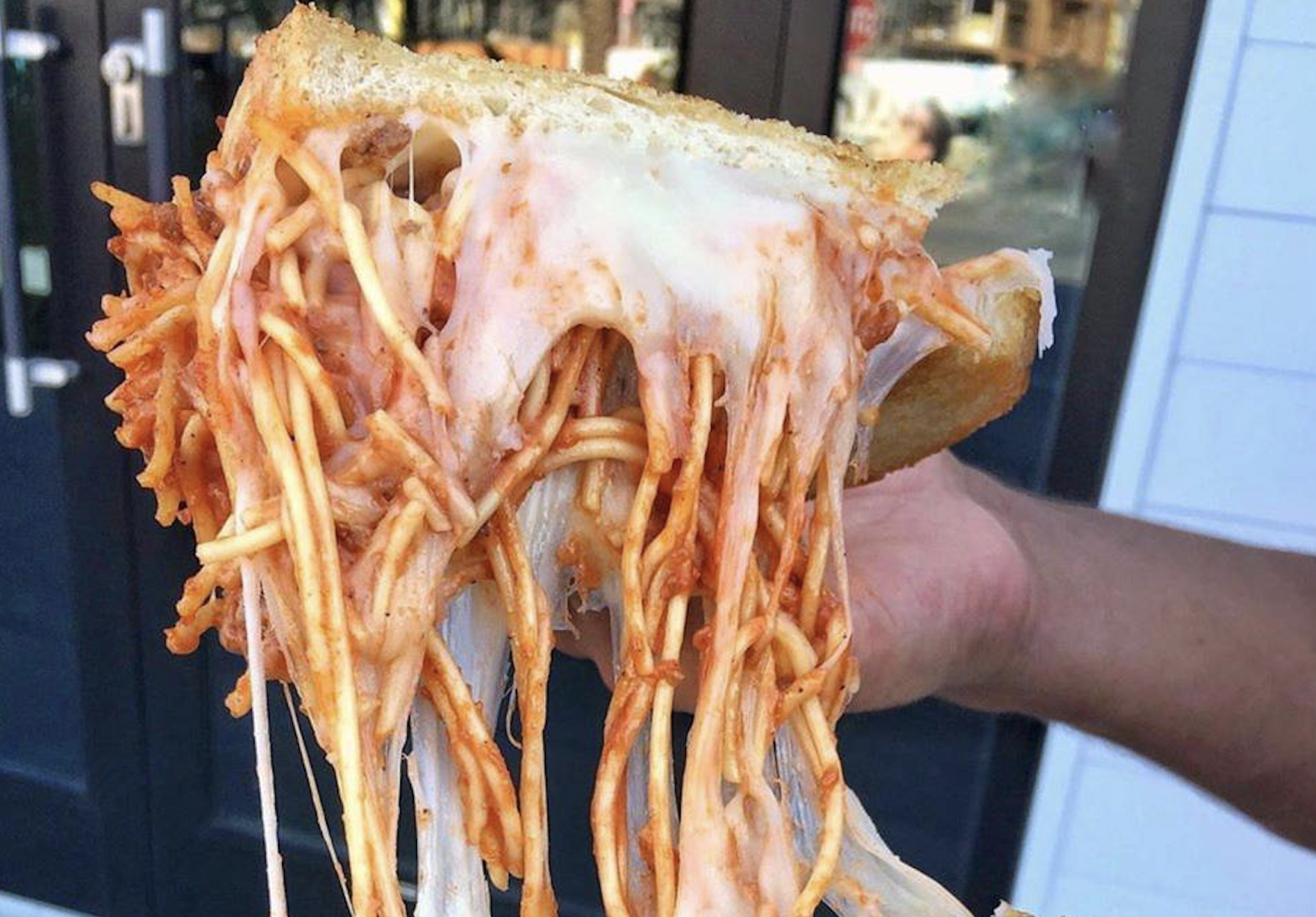 A hand holds a sandwich filled with spaghetti and melted cheese, with cheese stretching as the sandwich is pulled apart. The background shows a building with large glass doors.