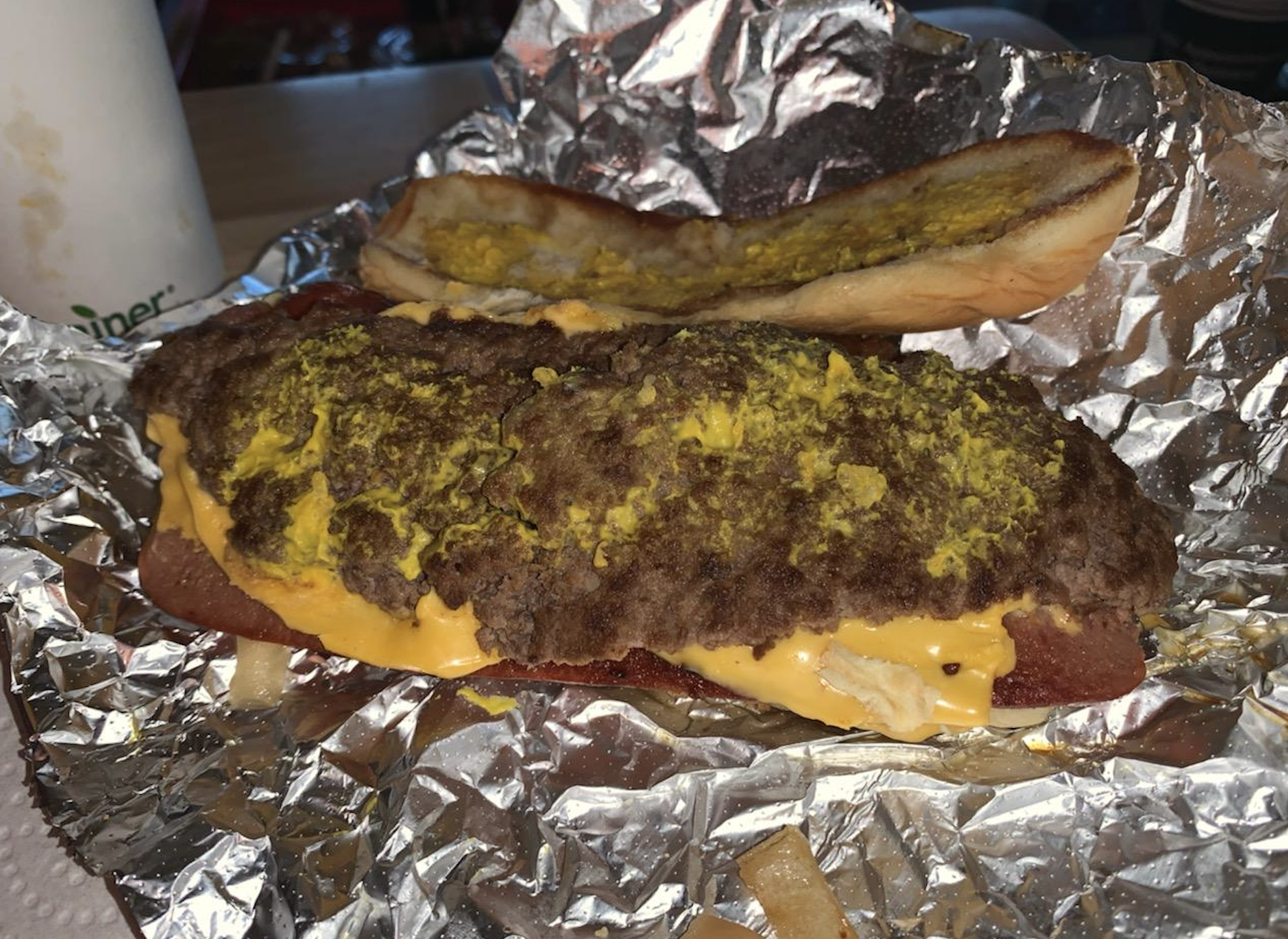 A sandwich on foil with a beef patty, melted cheese, mustard, and a split hoagie bun. Some fries are visible nearby. The sandwich appears messy and the top bun is off to the side.