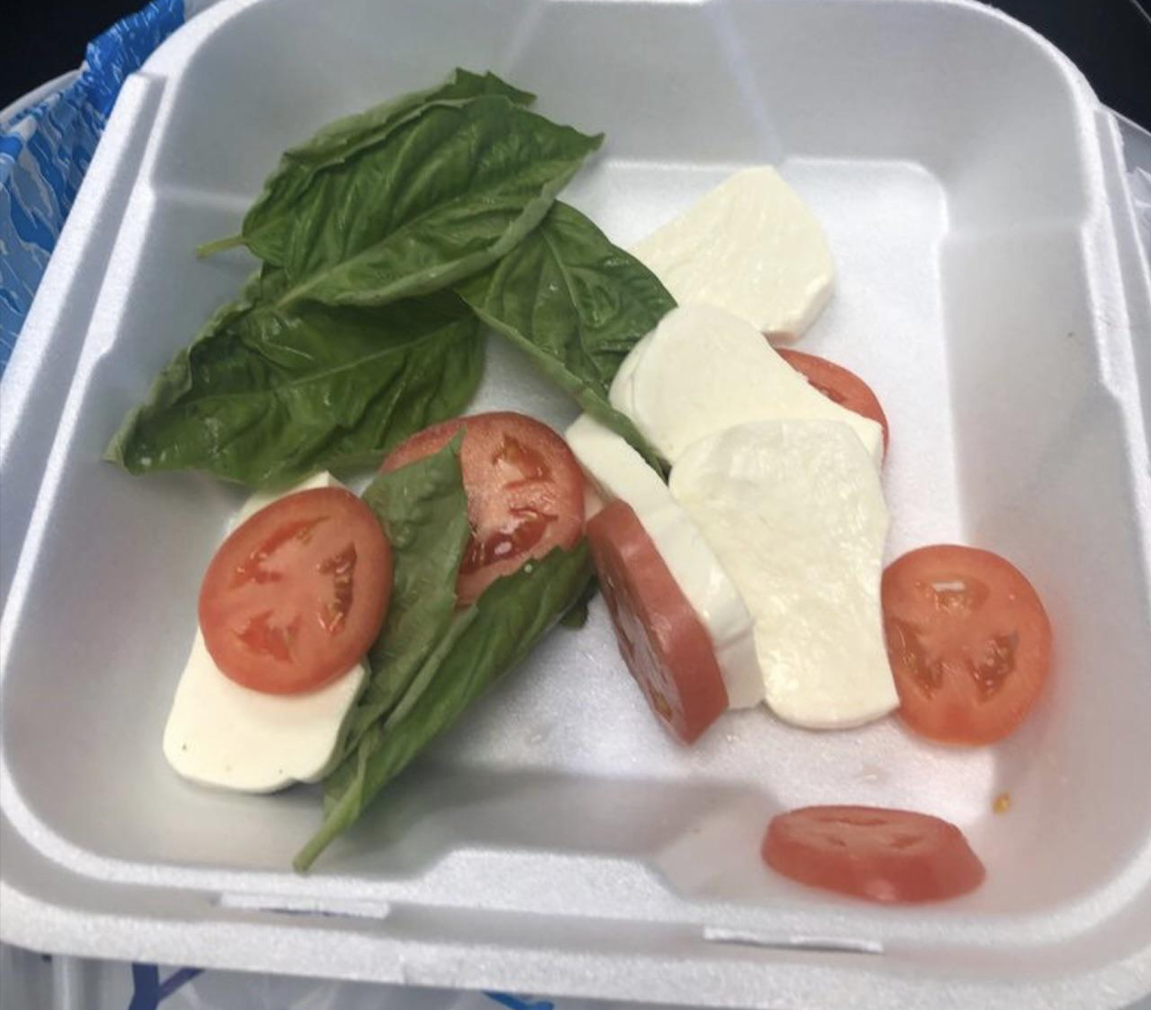 Sliced mozzarella, tomato, and basil leaves arranged in a white foam takeout container with a blue plastic utensil partially visible on the side.