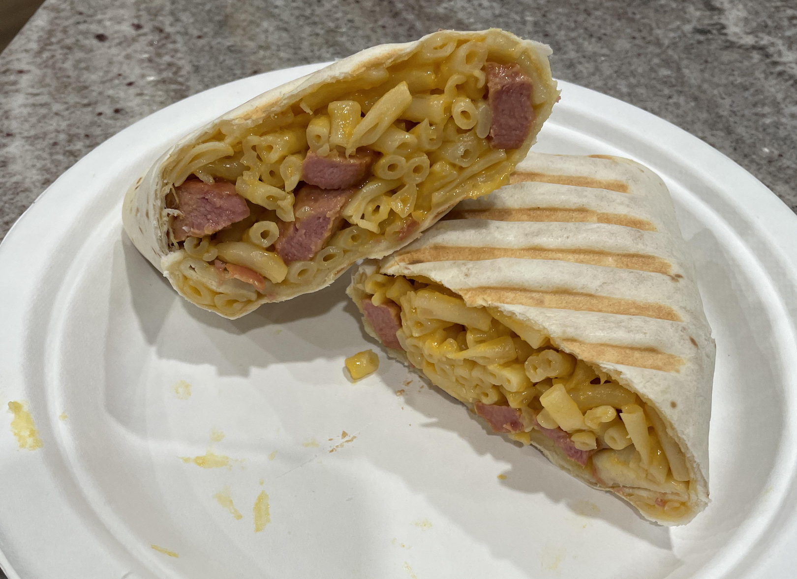A grilled wrap cut in half reveals macaroni and cheese with diced sausage inside, served on a white paper plate on a speckled countertop.