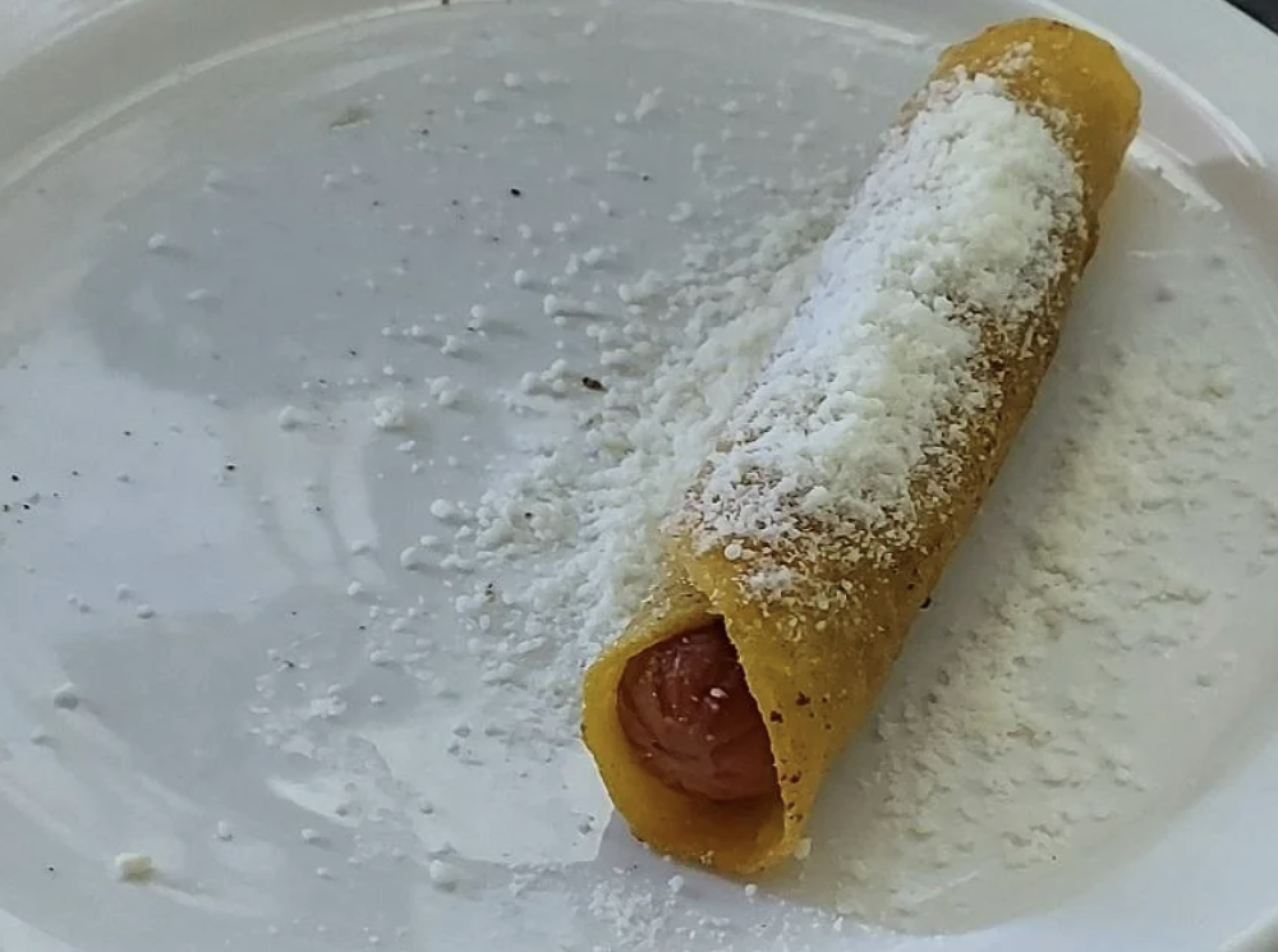 A rolled yellow corn tortilla with a sausage inside, topped with white grated cheese, sits on a white plate with cheese scattered around.