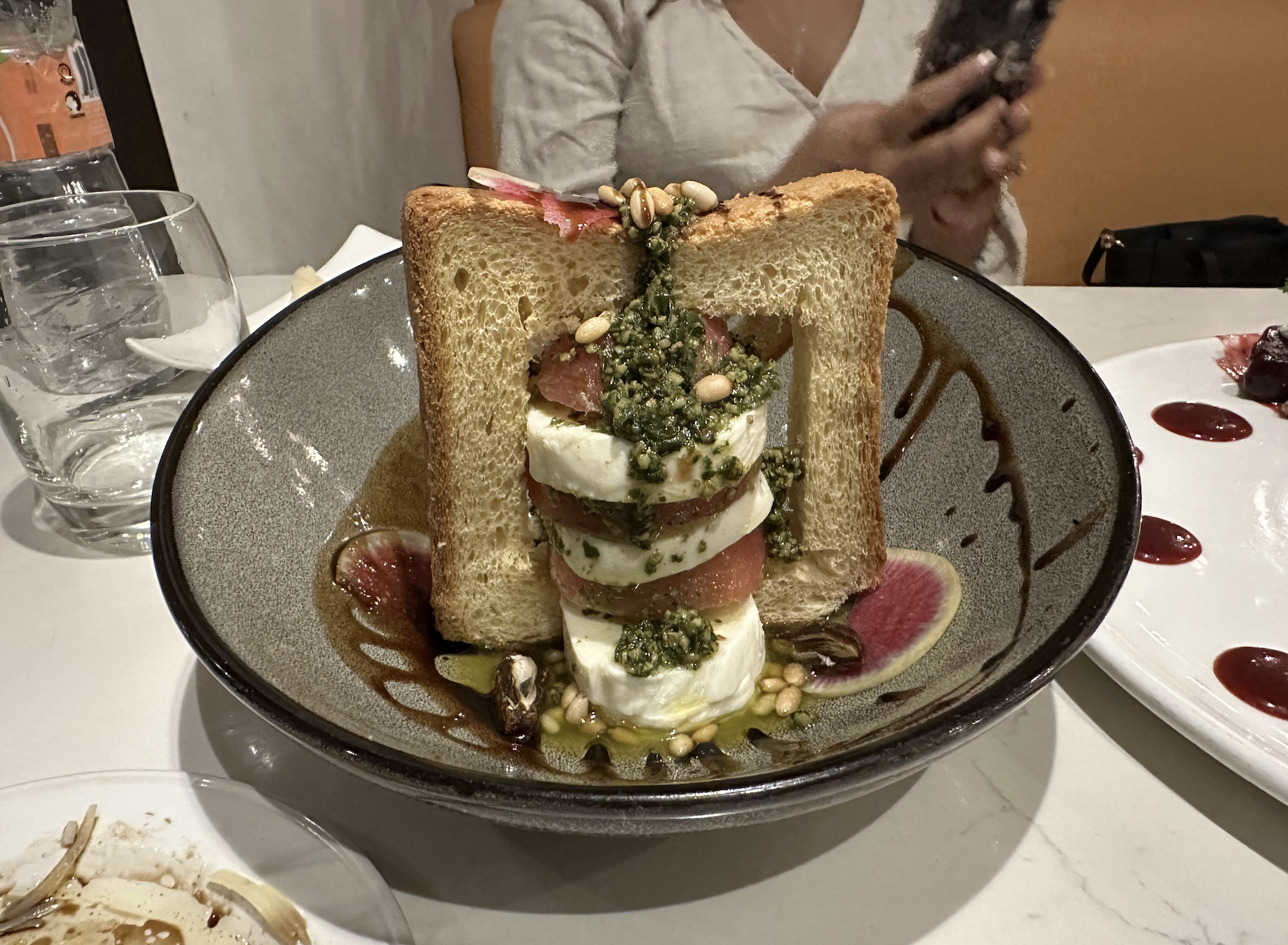 A plated dish features stacked mozzarella, tomato, and pesto, framed by two slices of toasted bread. Pine nuts and oil garnish the plate. A seated person holds a phone in the blurred background.