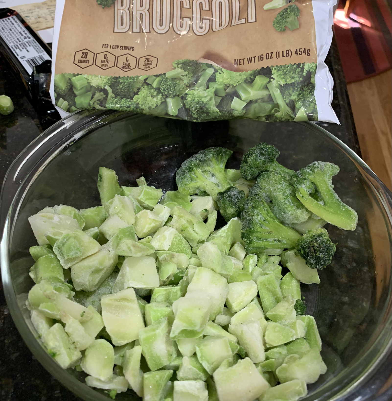 A glass bowl with frozen broccoli pieces, some florets and some stems, sits on a counter. Behind it is an open bag labeled “Broccoli.” The setting appears to be a kitchen with visible countertop and rug.