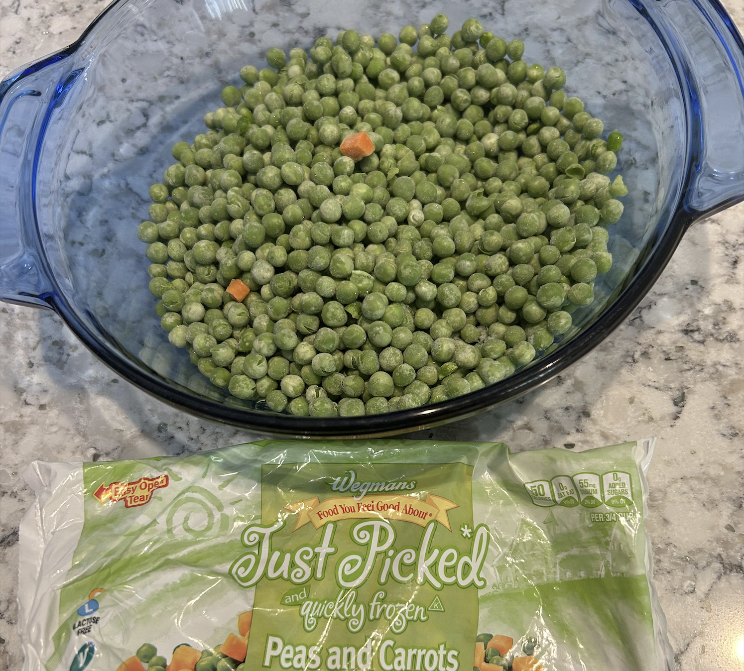A glass dish filled with mostly green peas and a few carrot pieces sits on a marble counter. Below it is an opened bag labeled “Just Picked Peas and Carrots.”