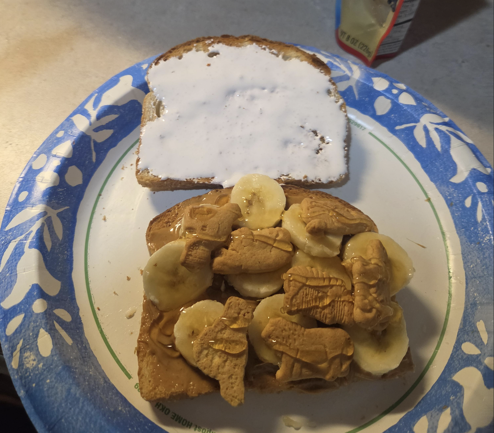 A sandwich on a paper plate with one slice spread with marshmallow fluff. The other slice has peanut butter, banana slices, honey, and animal-shaped graham crackers—an inventive creation that might just be one of the most delightful food fails you’ve ever tasted.