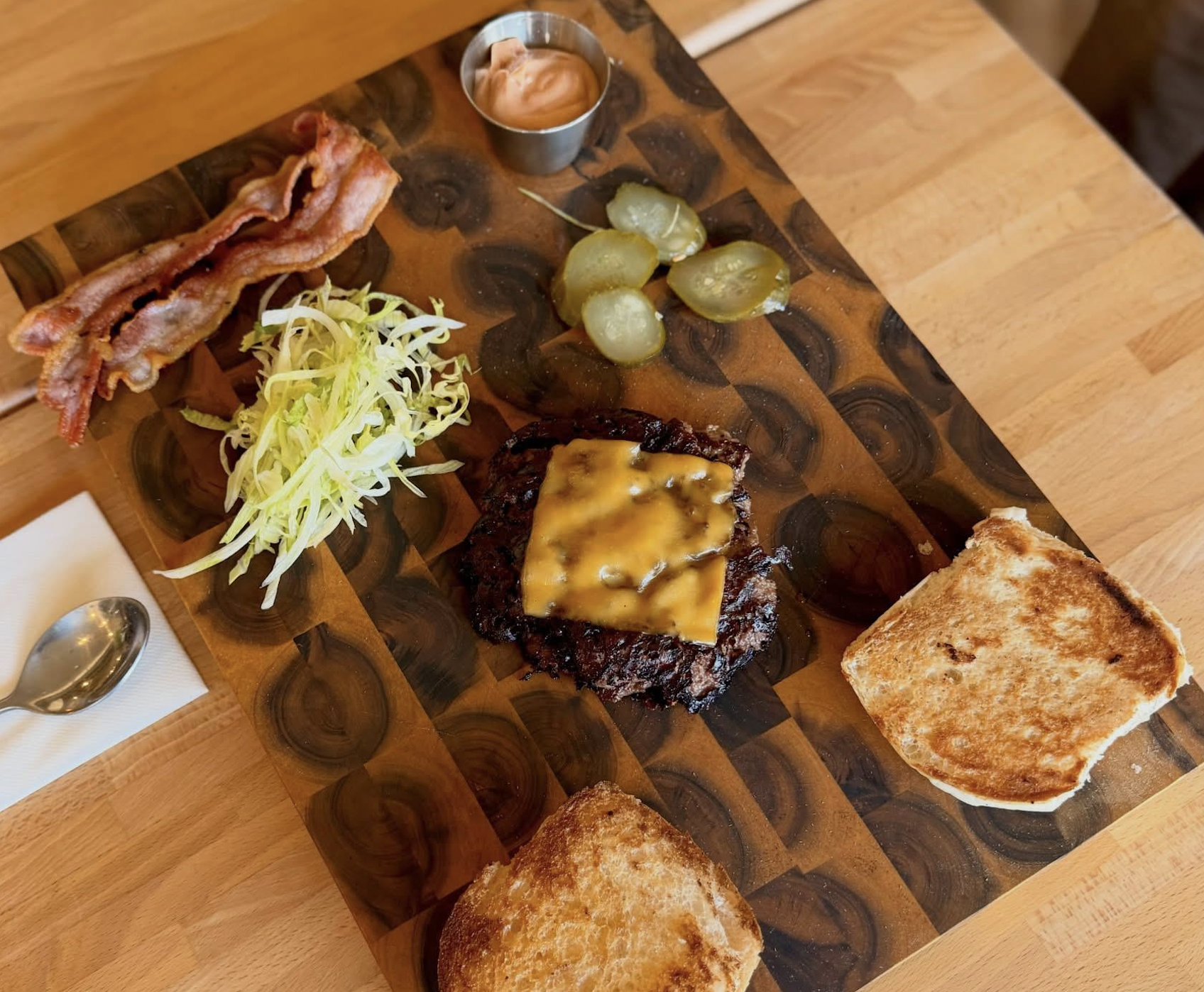 Burger ingredients on a wooden board: cooked bacon strips, shredded lettuce, grilled beef patty with melted cheese, pickles, toasted buns, and a small cup of sauce—everything you need to avoid food fails and build the perfect burger. A spoon and napkin are beside the board.