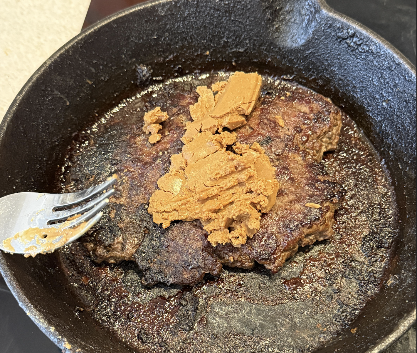 A cooked beef patty in a cast iron skillet topped with a chunk of peanut butter, with a fork resting on the left side—a creative combination that could easily earn a spot among memorable food fails.