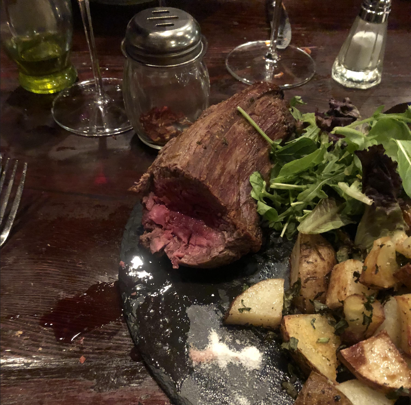 A plate with rare steak, roasted potatoes, and mixed greens on a dark wooden table, surrounded by wine glasses, a bottle of olive oil, and salt and pepper shakers.