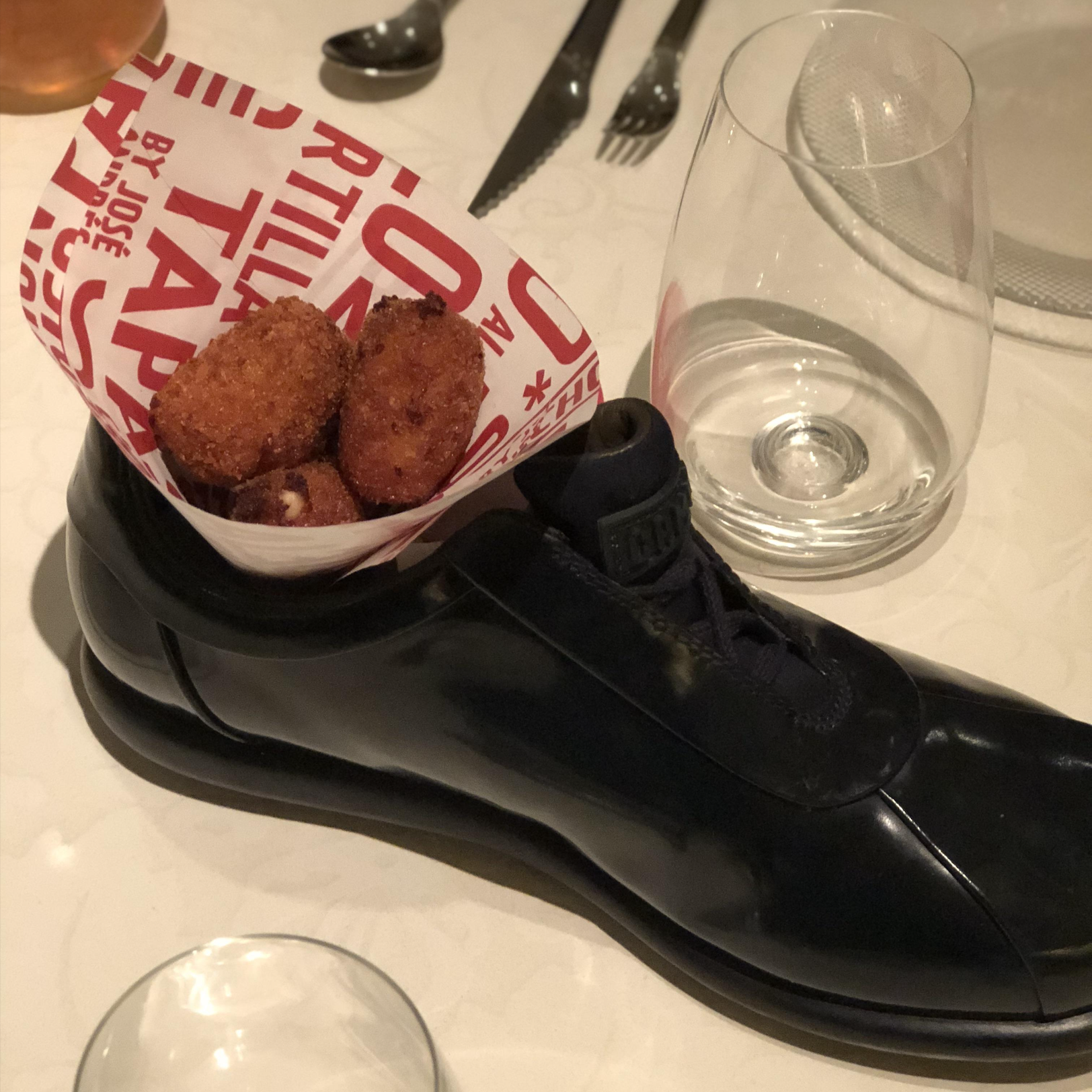 A black dress shoe holds fried croquettes in a red and white paper cone, placed on a table set with cutlery, a glass, and a plate.