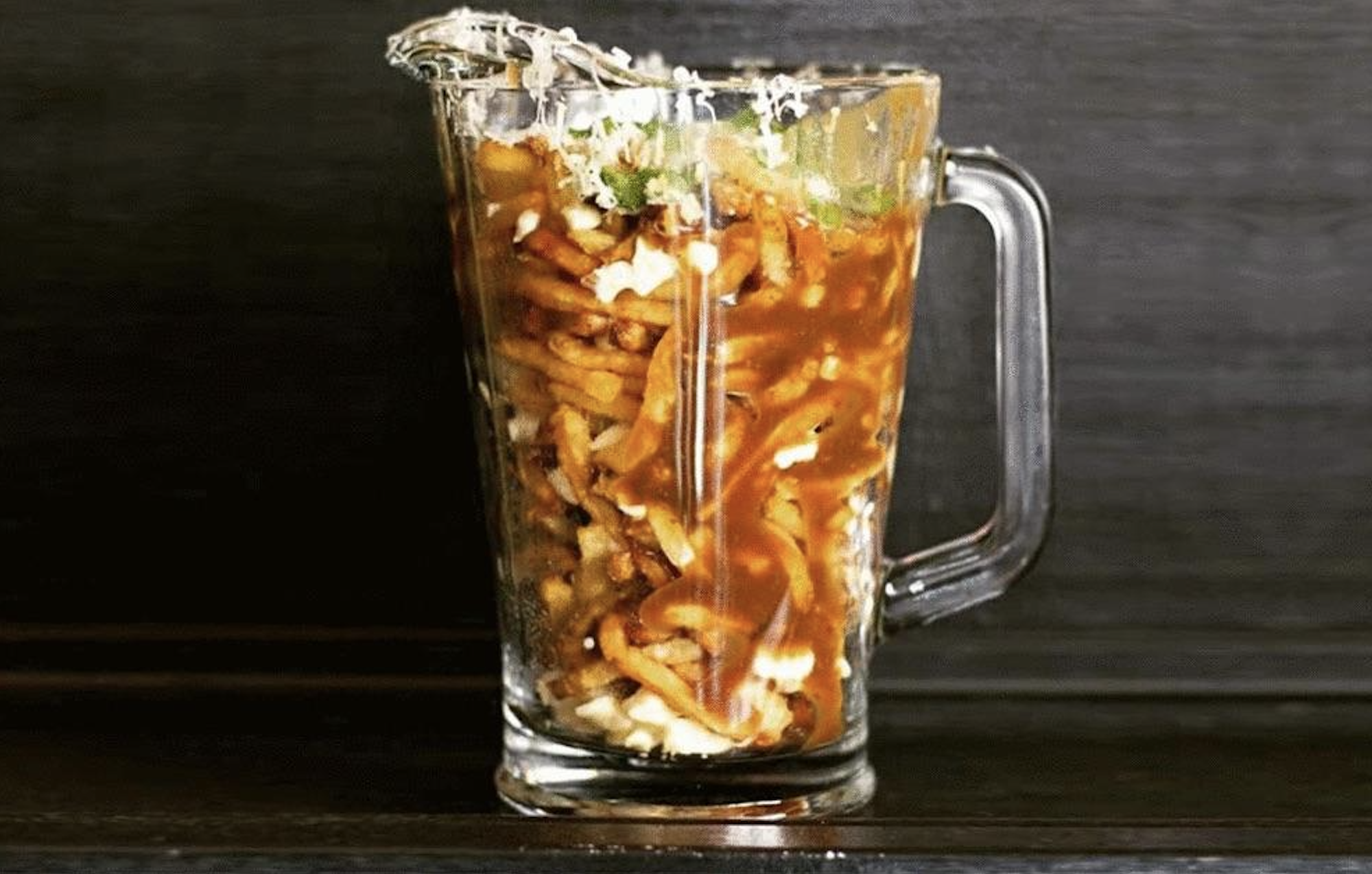 A glass pitcher filled with poutine, featuring layers of french fries, cheese curds, gravy, and green onions, against a dark background.