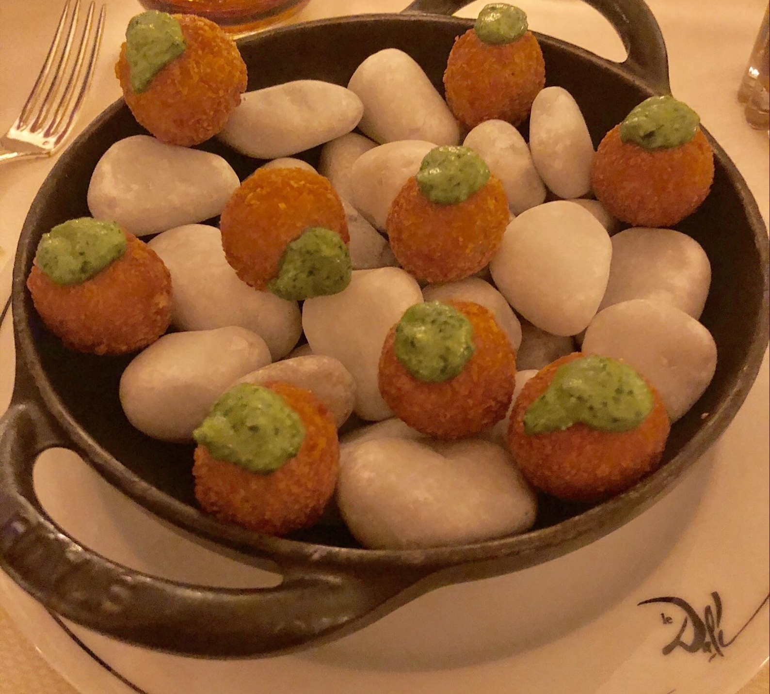 A black dish holds white smooth stones topped with round fried bites and a dollop of green sauce, arranged artistically.