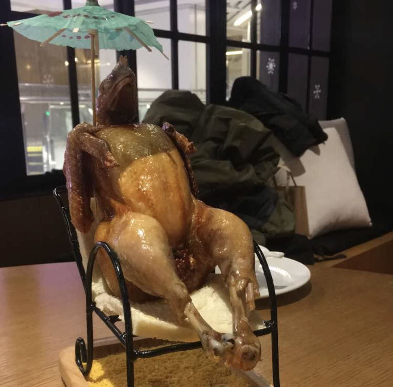 A roasted chicken posed lounging on a mini chair with a cocktail umbrella over its head, mimicking a person relaxing on vacation.