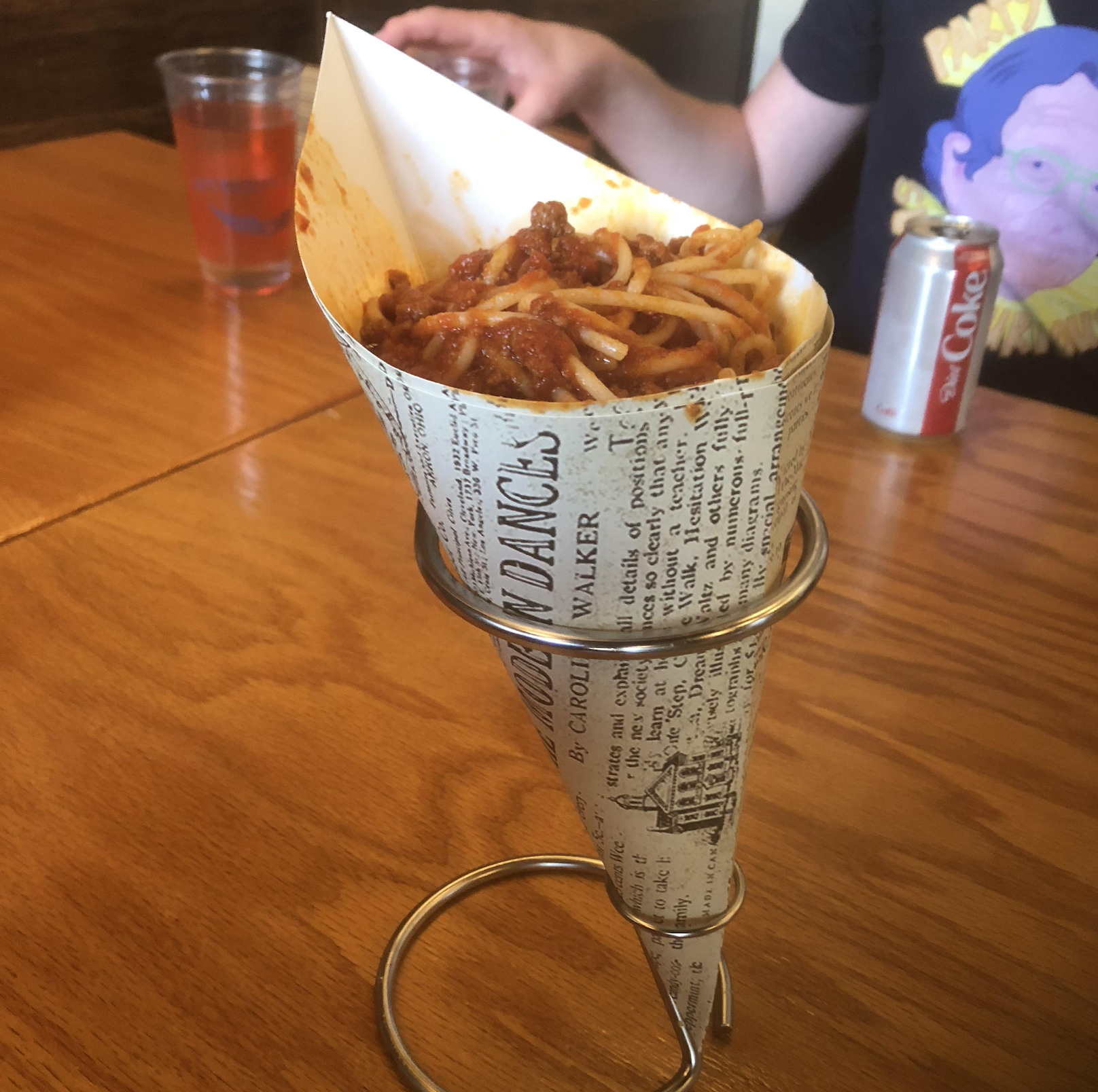 Spaghetti with sauce served in a paper cone, held upright in a metal stand on a wooden table. A hand, a drink, and a person wearing a colorful t-shirt are visible in the background.