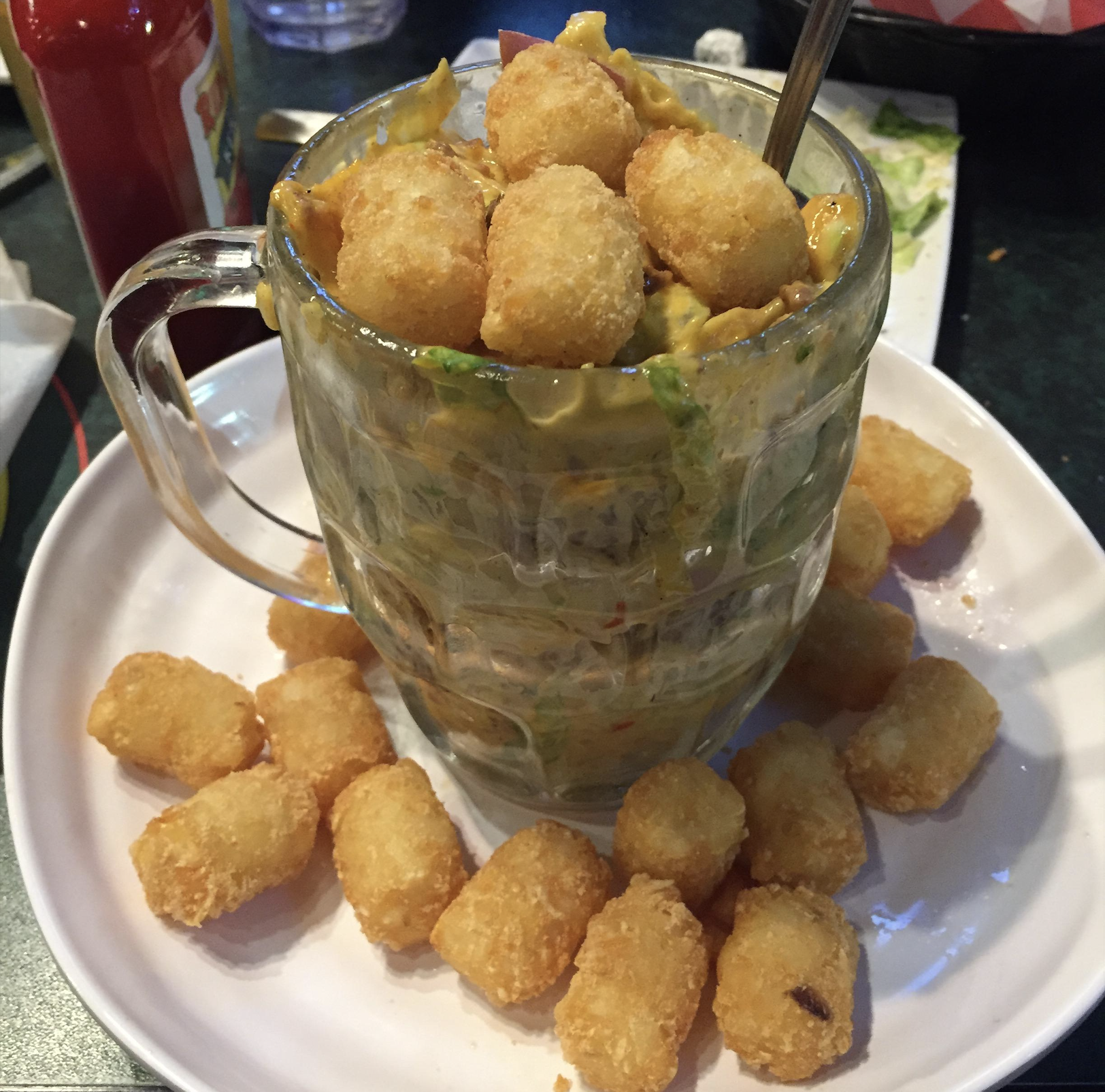 A glass mug filled with salad and topped with tater tots, placed on a white plate with more tater tots scattered around the mug.