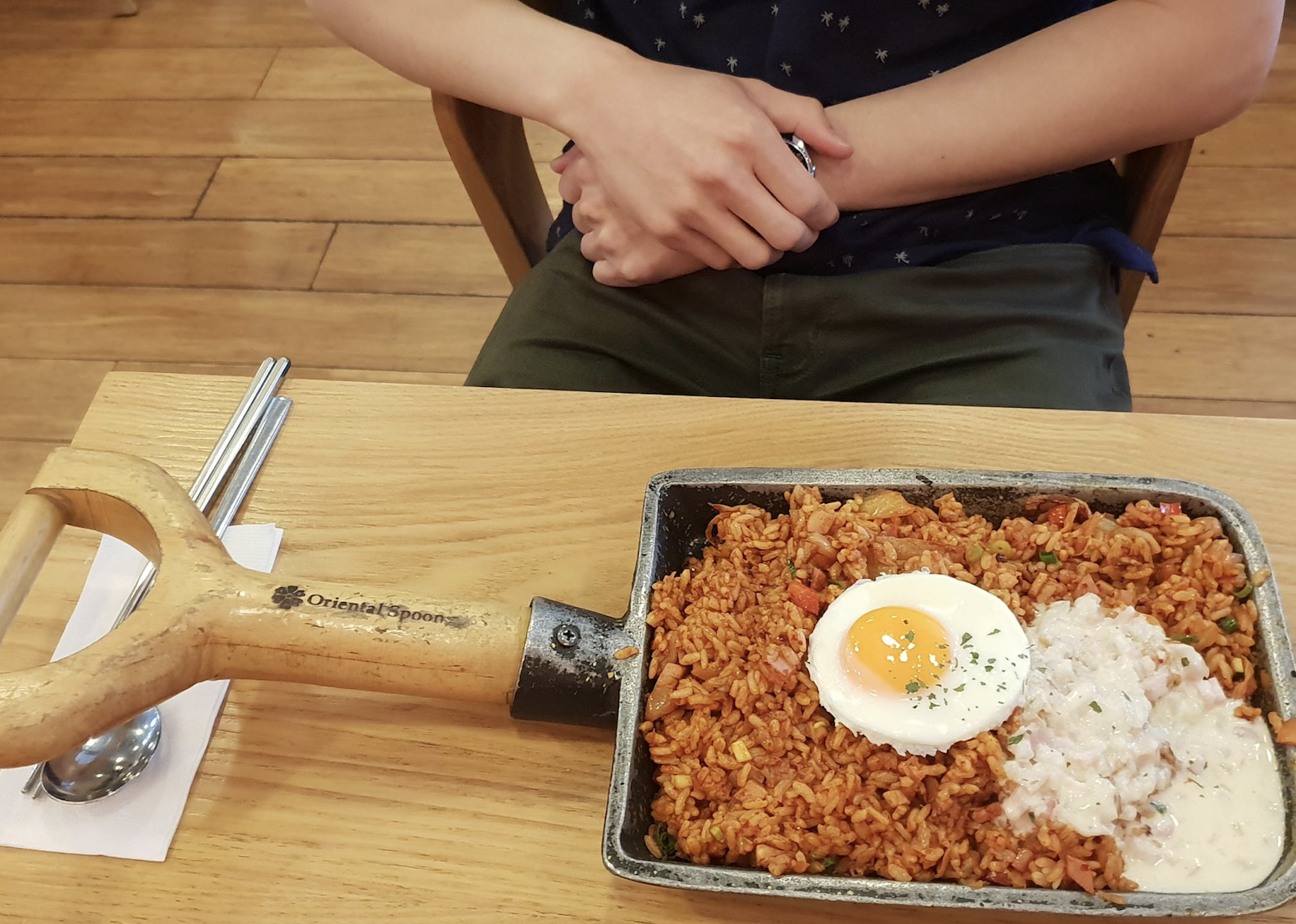 A person sits at a table with arms folded. In front is a skillet of fried rice topped with a sunny-side-up egg and creamy sauce. The skillet has a wooden handle and is served with chopsticks and a spoon on the side.