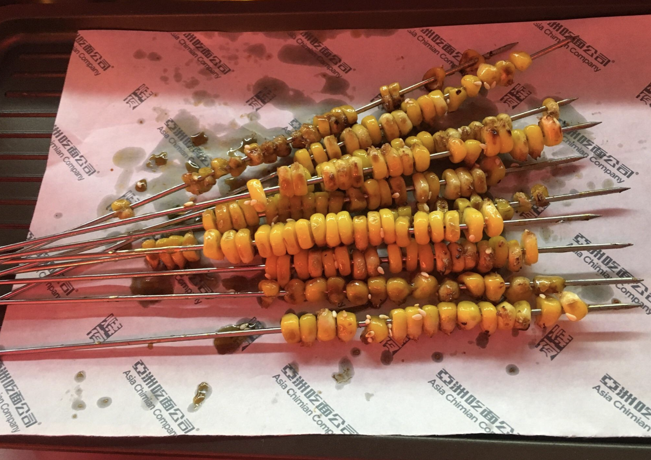 Several metal skewers threaded with grilled corn kernels rest on a paper-lined tray, with some seasoning and oil visible. The paper has Asian characters and English text printed on it.