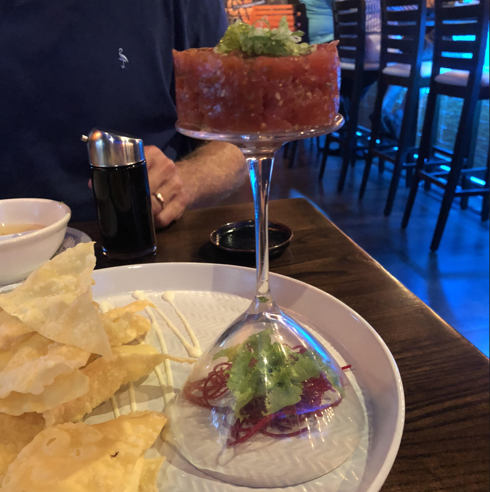 A glass filled with diced ahi tuna and garnished with green onions sits upside down on a plate with chips and shredded vegetables at a restaurant table.