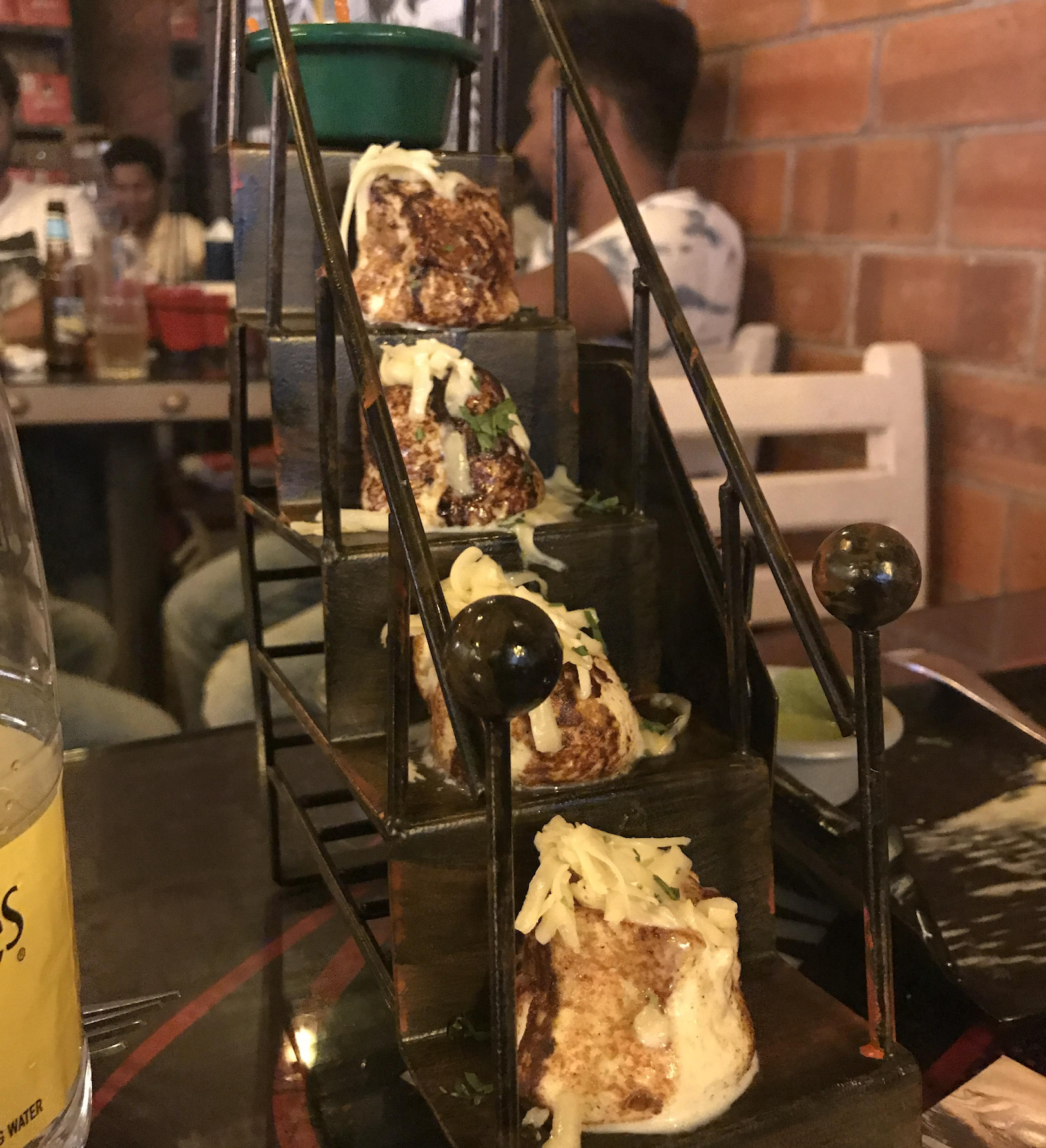 Four pieces of cheesy grilled food are displayed on a miniature staircase-shaped metal stand at a restaurant, with people and brick walls in the background.