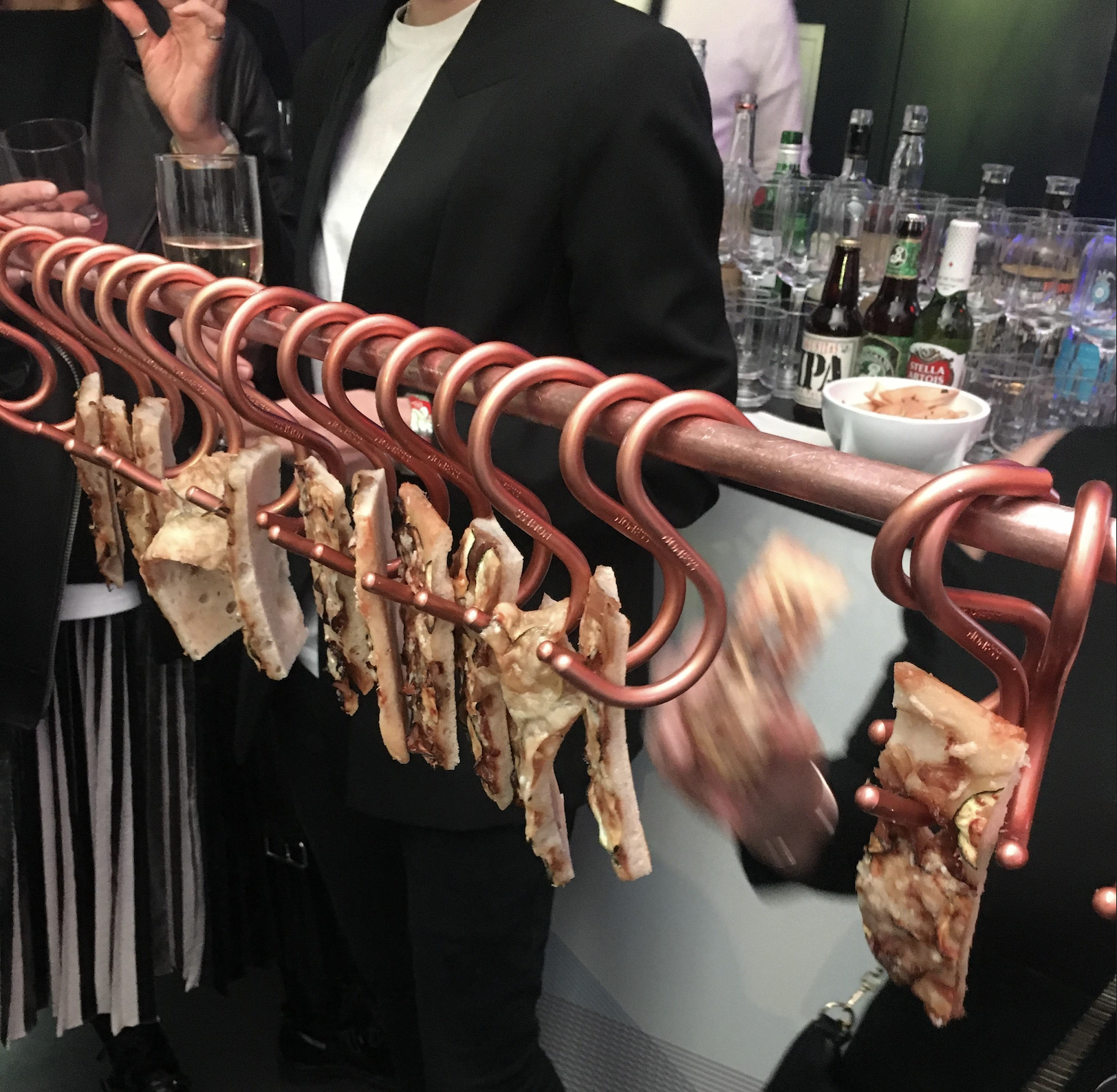 Several pieces of pizza are hanging on large metal hooks from a rod at a social gathering, with drinks and snacks visible in the background.