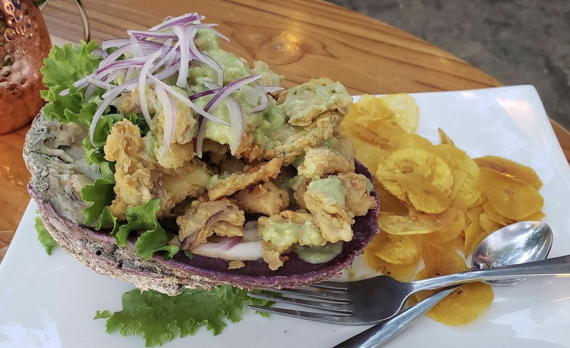 Fried clams topped with avocado salsa and red onions served in a shell, with plantain chips on the side, on a white plate with a fork and spoon.