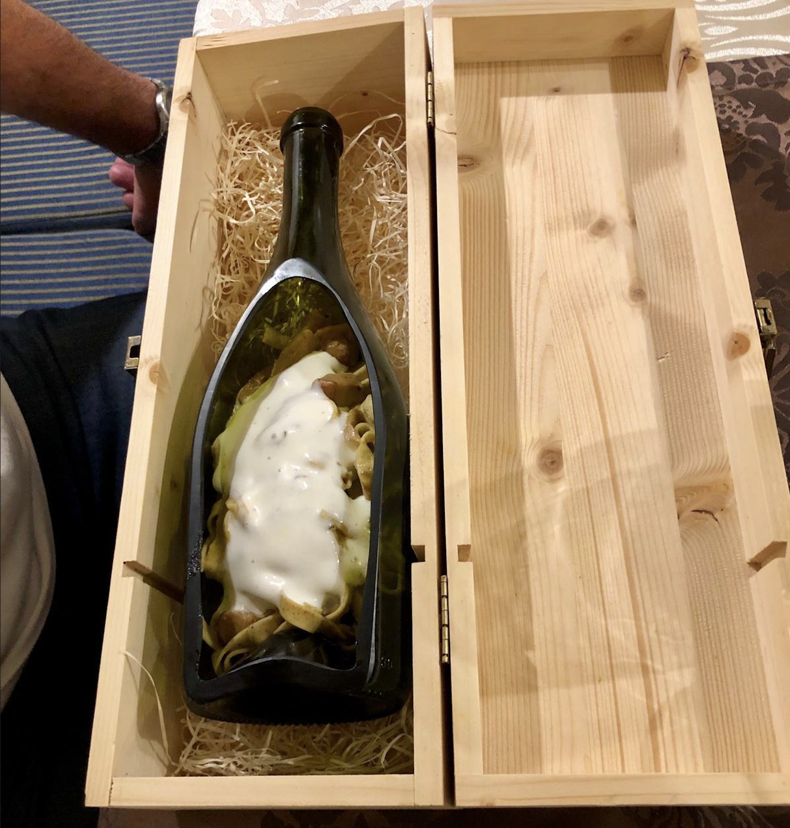 A cut wine bottle filled with pasta and sauce is placed in a wooden box lined with straw. A person’s arm rests at the edge of the image.