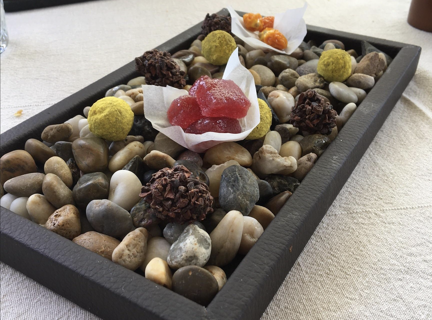 Assorted colorful candies and chocolate truffles are served on a bed of smooth pebbles in a rectangular tray lined with paper.