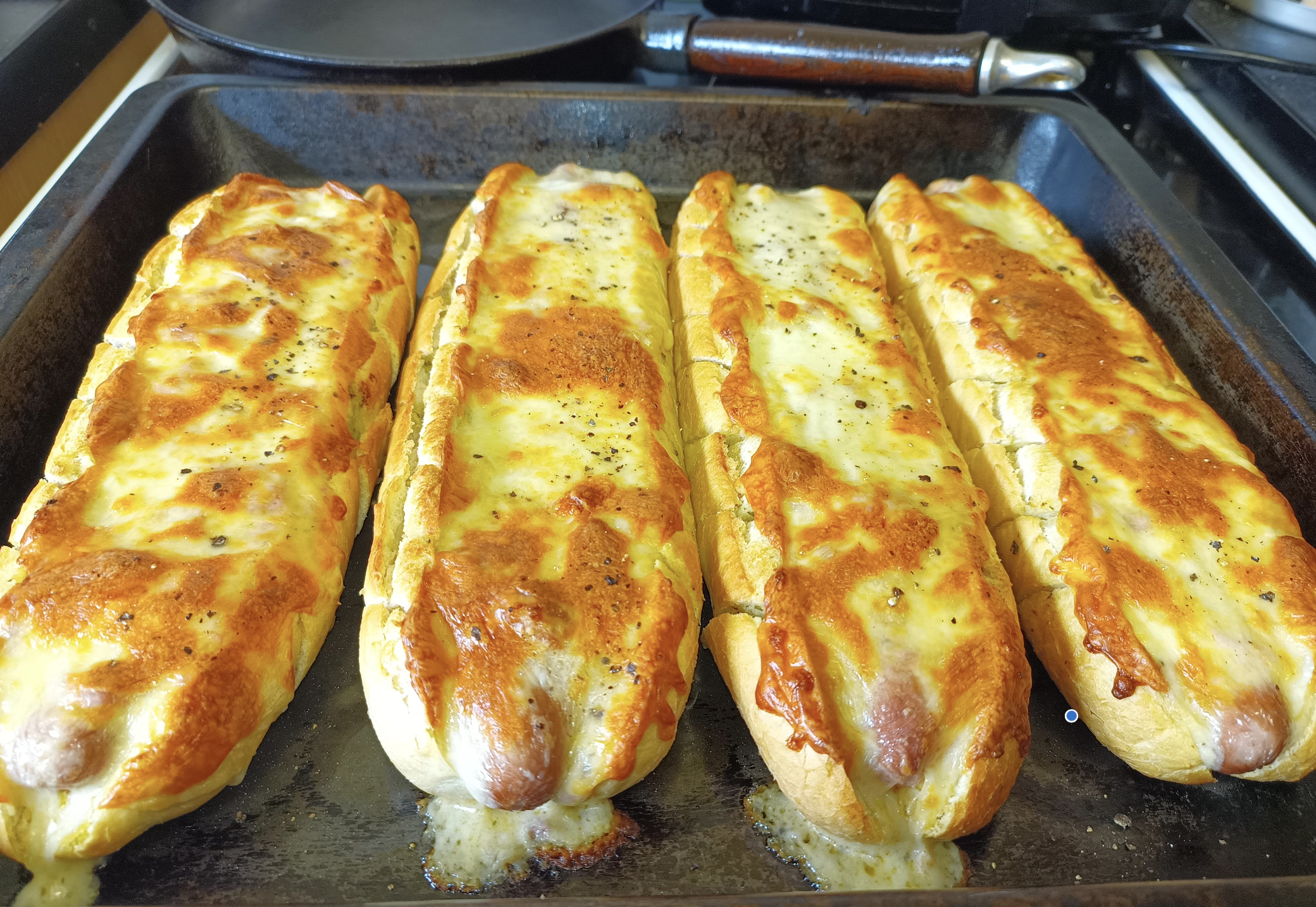 Four baked cheese-topped baguettes on a dark oven tray, each split open and filled with melted cheese, with golden brown tops and some herbs sprinkled on them.