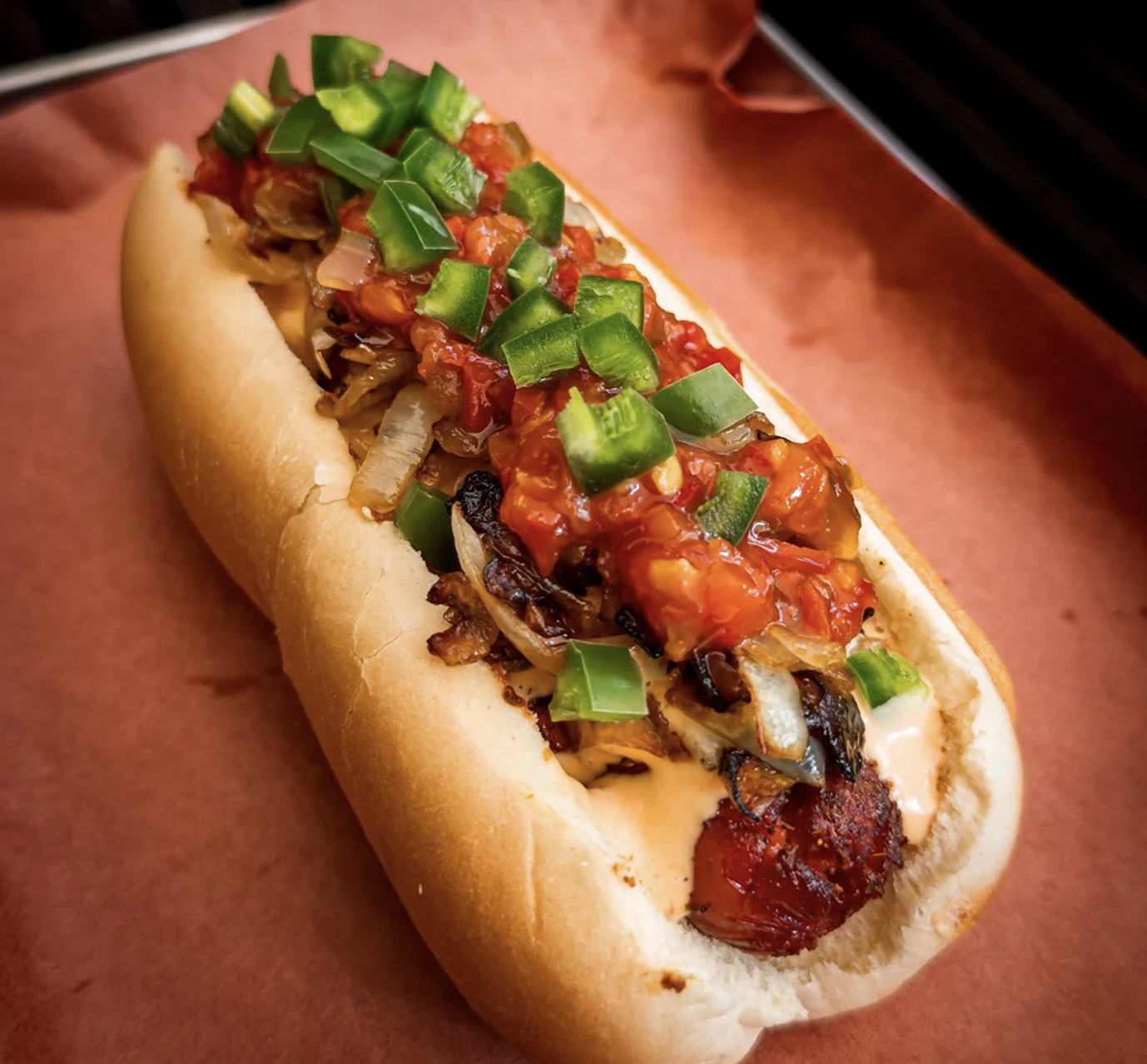 A grilled sausage in a bun topped with sautéed onions, diced jalapeños, and chunky salsa, resting on brown parchment paper.