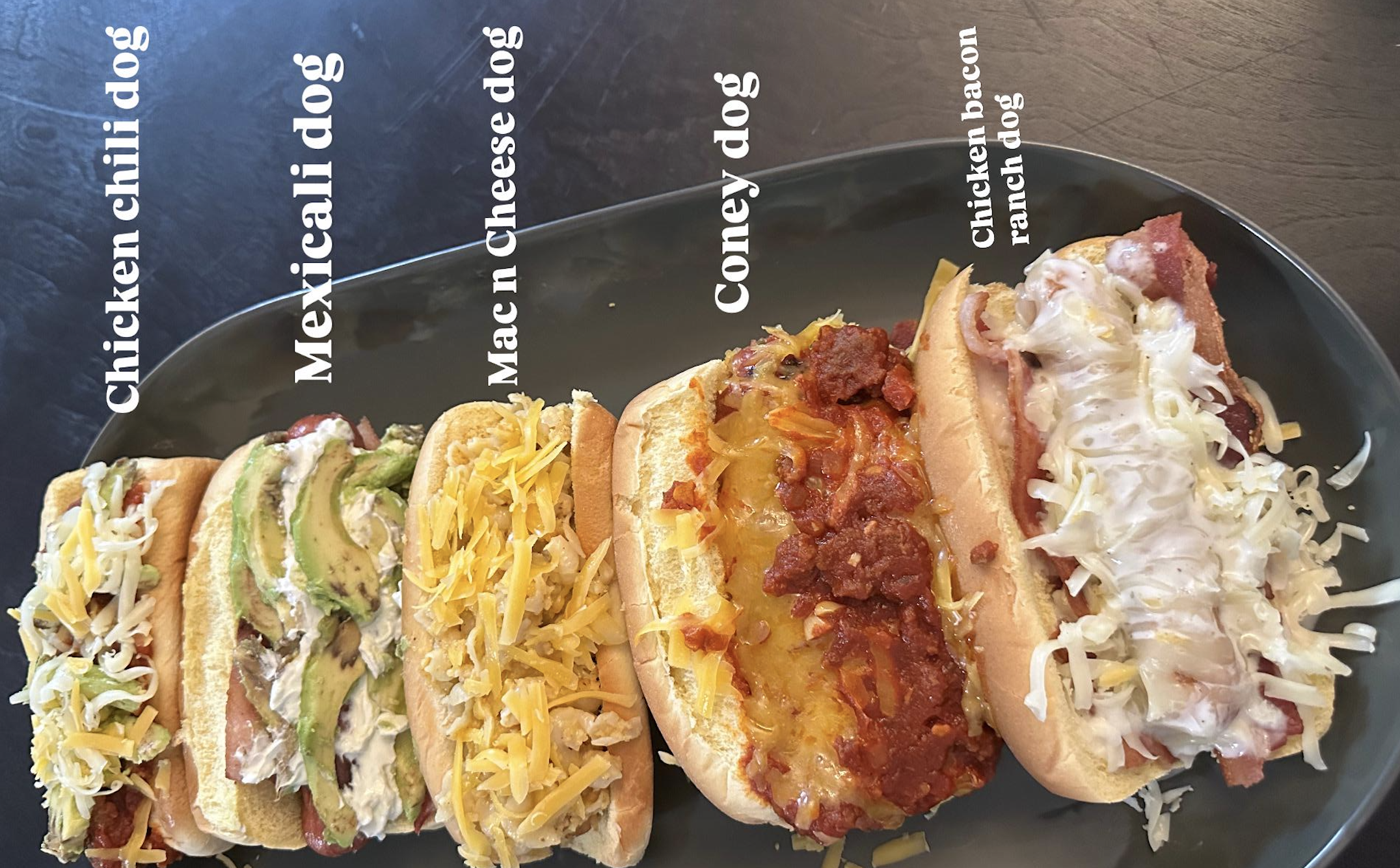 Five hot dogs on a black platter, each labeled: Chicken chili dog, Mexicali dog, Mac n Cheese dog, Coney dog, and Chicken bacon ranch dog, each with different toppings and melted cheese.